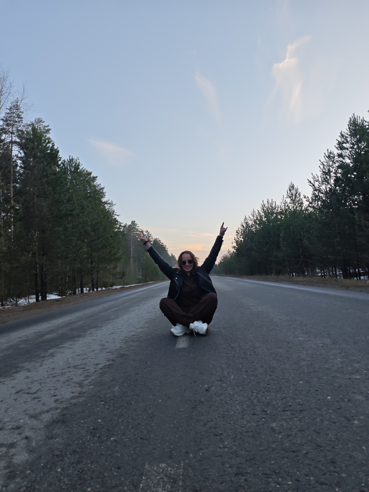A woman is sitting cross-legged on an asphalt road with trees on both sides, raising her arms above her head. She is wearing sunglasses, a dark jacket, a brown jumpsuit, and white sneakers. The road has some patches of white paint and appears to be in a rural area. The sky is a light blue with a touch of orange on the horizon. It is likely near Chaikovsky, Russia given the request.