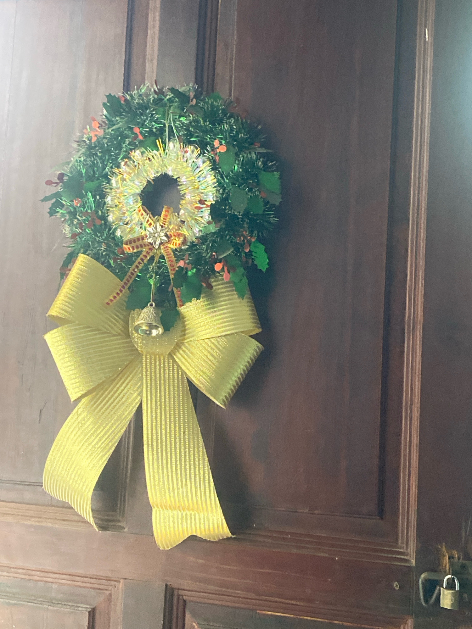 A Christmas wreath with a large yellow bow and a small bell hangs on a dark brown wooden door. The wreath is made of green tinsel and has small red berries. A shiny gold ornament is in the center of the wreath. The bow is made of a ribbed yellow ribbon. The door has paneled sections. A small padlock is visible on the right side of the doorframe. The scene appears to be a domestic setting in Batam, Indonesia.