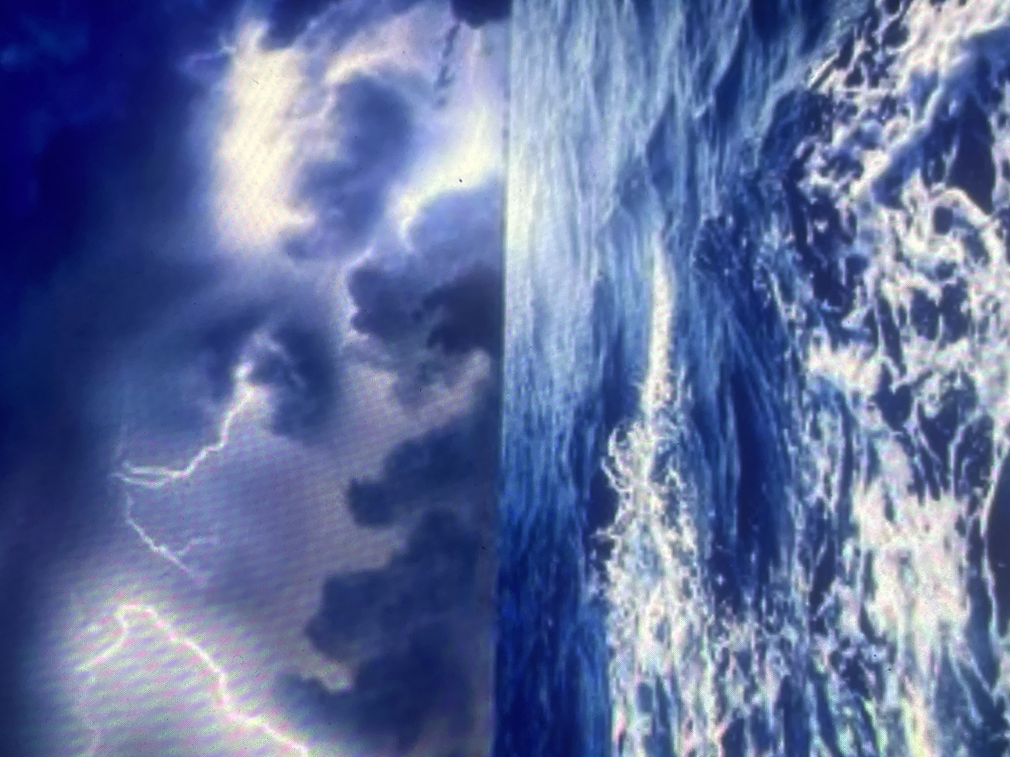 The scene depicts a dramatic and turbulent seascape under a severe thunderstorm, located off the coast of Zelenyi Hai, Ukraine. The upper half of the image is dominated by a dark, heavy sky filled with voluminous storm clouds in shades of deep indigo, grey, and purplish-blue. Multiple bright, jagged lightning strikes illuminate these formations, casting brilliant white and magenta-tinged light across the cloudscape, notably on the left and right sides of the frame. The horizon line is visible as a faint, indistinct demarcation separating the stormy sky from the ocean. The lower half of the image features a rough, agitated sea, with dark blue water churned into numerous whitecaps and foamy crests, particularly prominent in the foreground. The water's surface reflects the intense atmospheric light, highlighting the dynamic movement of the waves. No human subjects or man-made objects are visible within the frame. The environment is one of an open, stormy ocean.