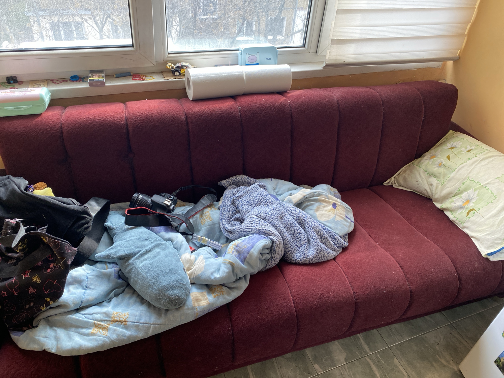 The image shows a messy, red sofa with a pillow on the right. There is a blue blanket, a plush shark toy, a camera, and bags on the left side of the sofa. A roll of paper towels sits on the sofa back, below the window. On the windowsill are small items and a container labeled "FINN." The location is most likely in a residential building and based on the presence of Bulgarian text in the image, the location is potentially in Sofia, Bulgaria. The scene depicts a cluttered living space. No people are visible.
