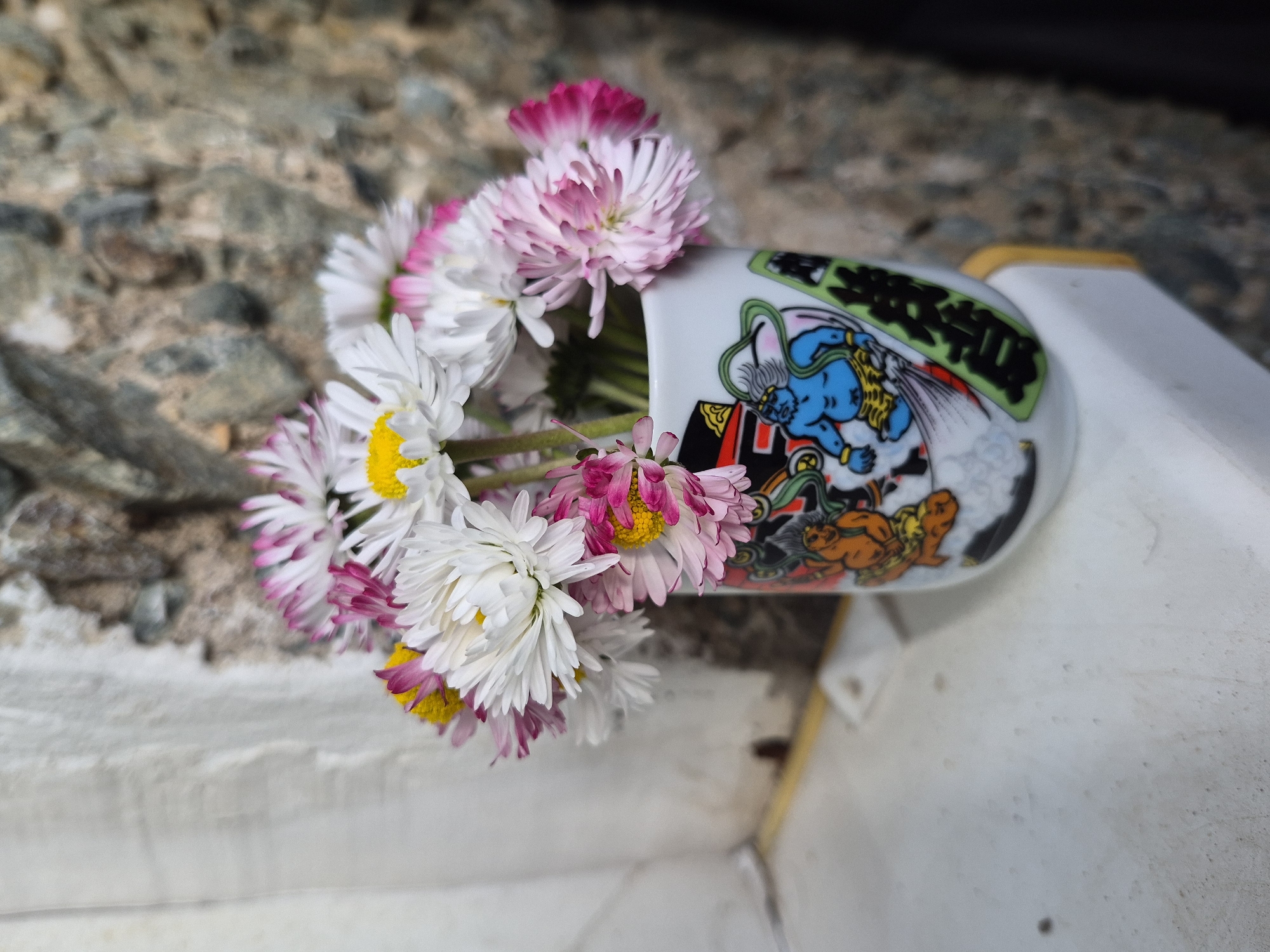 A still life features a small bouquet of daisies with white and pink-tipped petals and yellow centers, arranged in a Japanese-style teacup. The teacup has a white background and is adorned with a colorful illustration of a blue-skinned figure with Japanese text above. The teacup is positioned horizontally, allowing the flowers to spill out. The scene is set outdoors, with a weathered gray stone wall in the background and a partially visible white surface in the foreground. The location and specific details about the city are unknown.