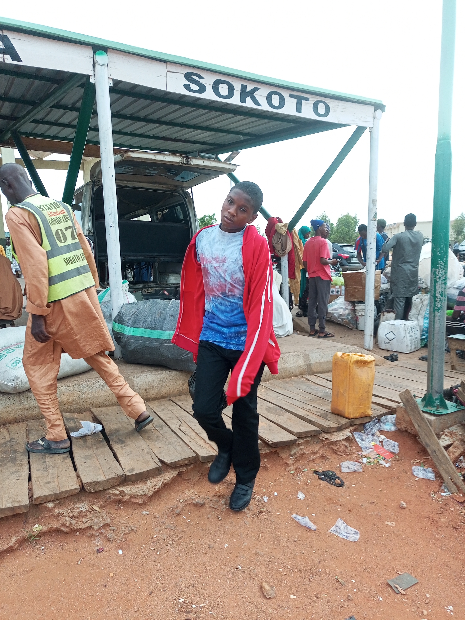The image depicts an outdoor scene at a transportation hub, possibly a bus or motor park, in Gombe, Nigeria. A sign overhead reads "SOKOTO," indicating a destination or route. The ground is a mix of reddish-brown earth and wooden planks that form a pathway. Several people are present, some appearing to be passengers or workers.
In the foreground, a young person in a red jacket over a blue and white tie-dye shirt and black pants is walking. To the left, another person wearing a tan outfit and a reflective vest with "STATION ATTENDANT" and "GOURS LIN" printed on it, along with the number "07", is walking. They are near a van with its trunk open, loaded with goods. Various bags, some appearing to be sacks of produce or merchandise, are scattered around. A bright yellow plastic container is visible on the wooden planks. The environment suggests a busy, possibly somewhat disorganized, area with luggage and cargo. There is litter on the ground. In the background, other individuals and vehicles are present, contributing to the atmosphere of a transit point.