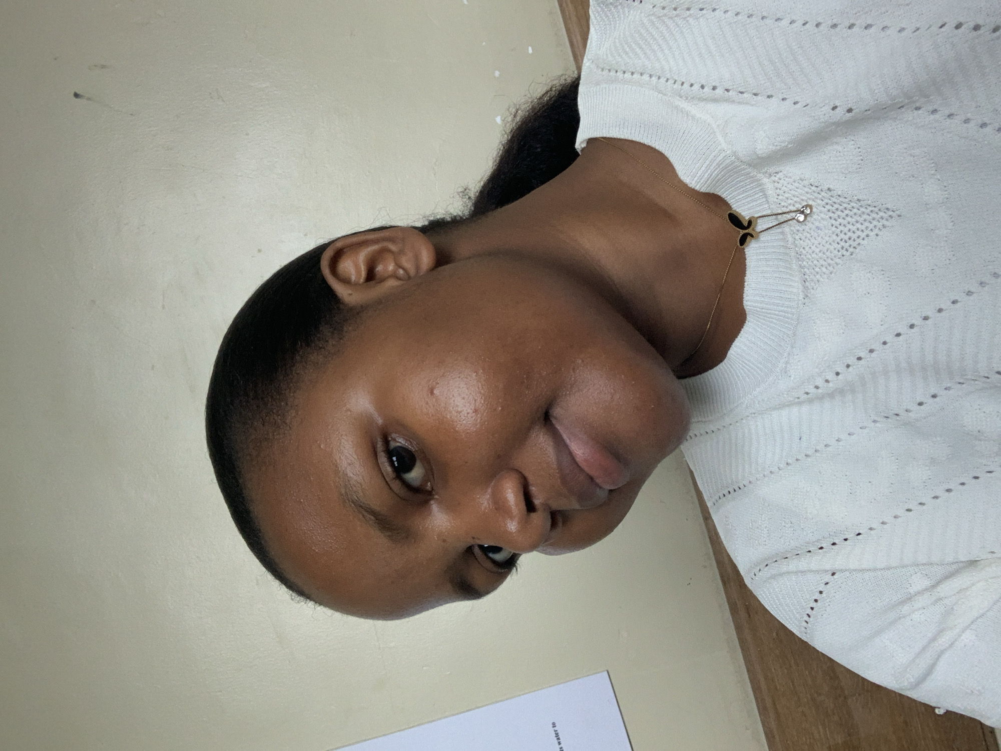 A young Black woman is positioned close to the camera. She has dark hair pulled back from her face, smooth brown skin, and is wearing a white knitted top. She appears to be wearing a necklace with a butterfly-shaped pendant. The background is a plain light beige or cream color. A sheet of paper with text partially visible at the bottom is present in the frame. There is no indication of location within the city of Dar es-Salaam, Tanzania.