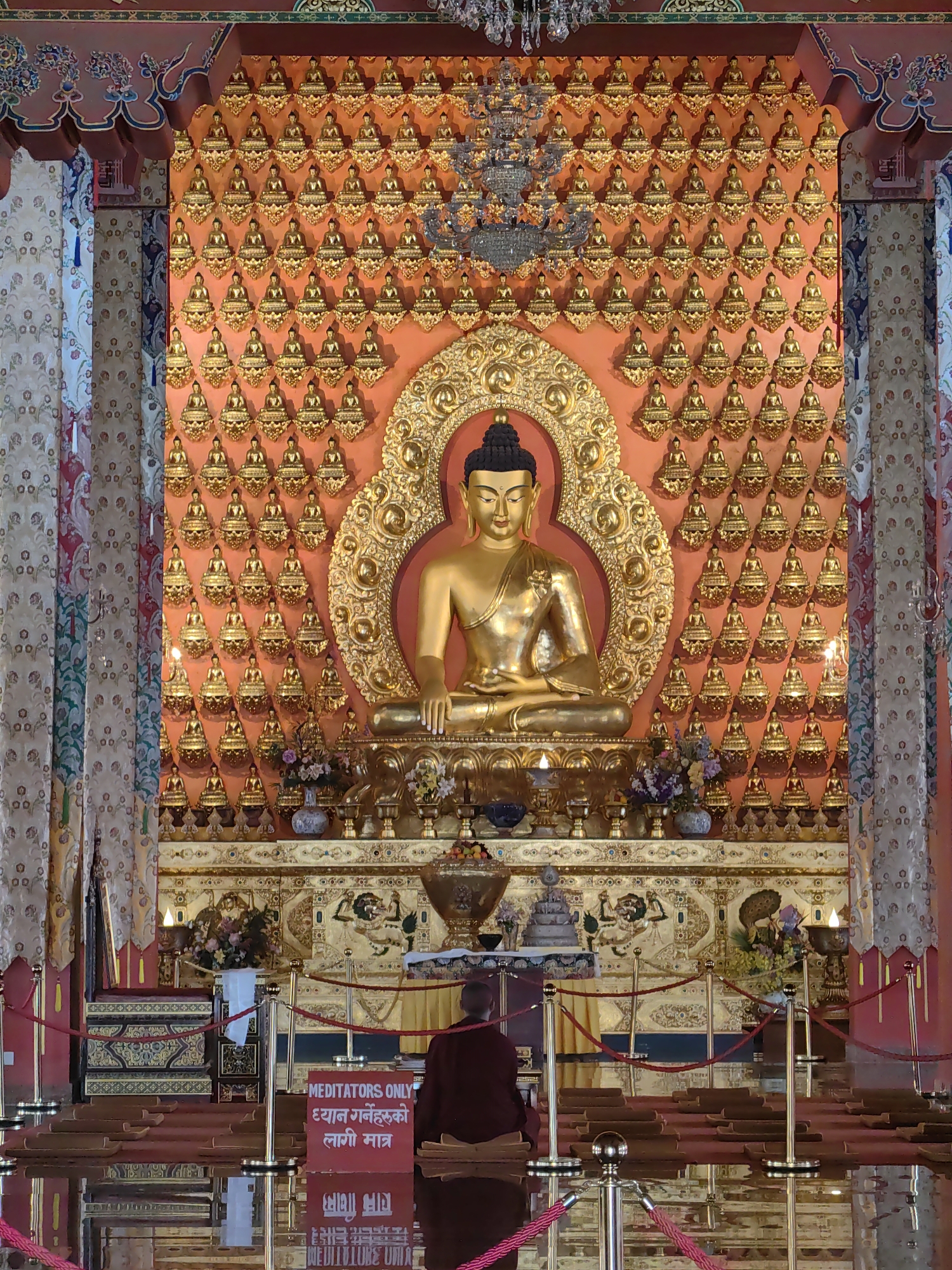 This image depicts the interior of a Buddhist temple in Parsa, Nepal. The central focus is a large, golden statue of Buddha seated in a meditative pose. Behind the main Buddha statue, the wall is adorned with a grid of numerous smaller golden Buddha statues. The decor is ornate, with gold accents and colorful drapery on the sides. In the foreground, a person in maroon robes is seated, facing the main Buddha statue, seemingly in meditation. The floor is highly reflective, mirroring the scene above. Red ropes and stanchions are placed to guide visitors and create a designated viewing area. A sign is visible with text in English and Nepali, indicating "MEDITATORS ONLY." The environment appears serene and sacred, characteristic of a place of worship.