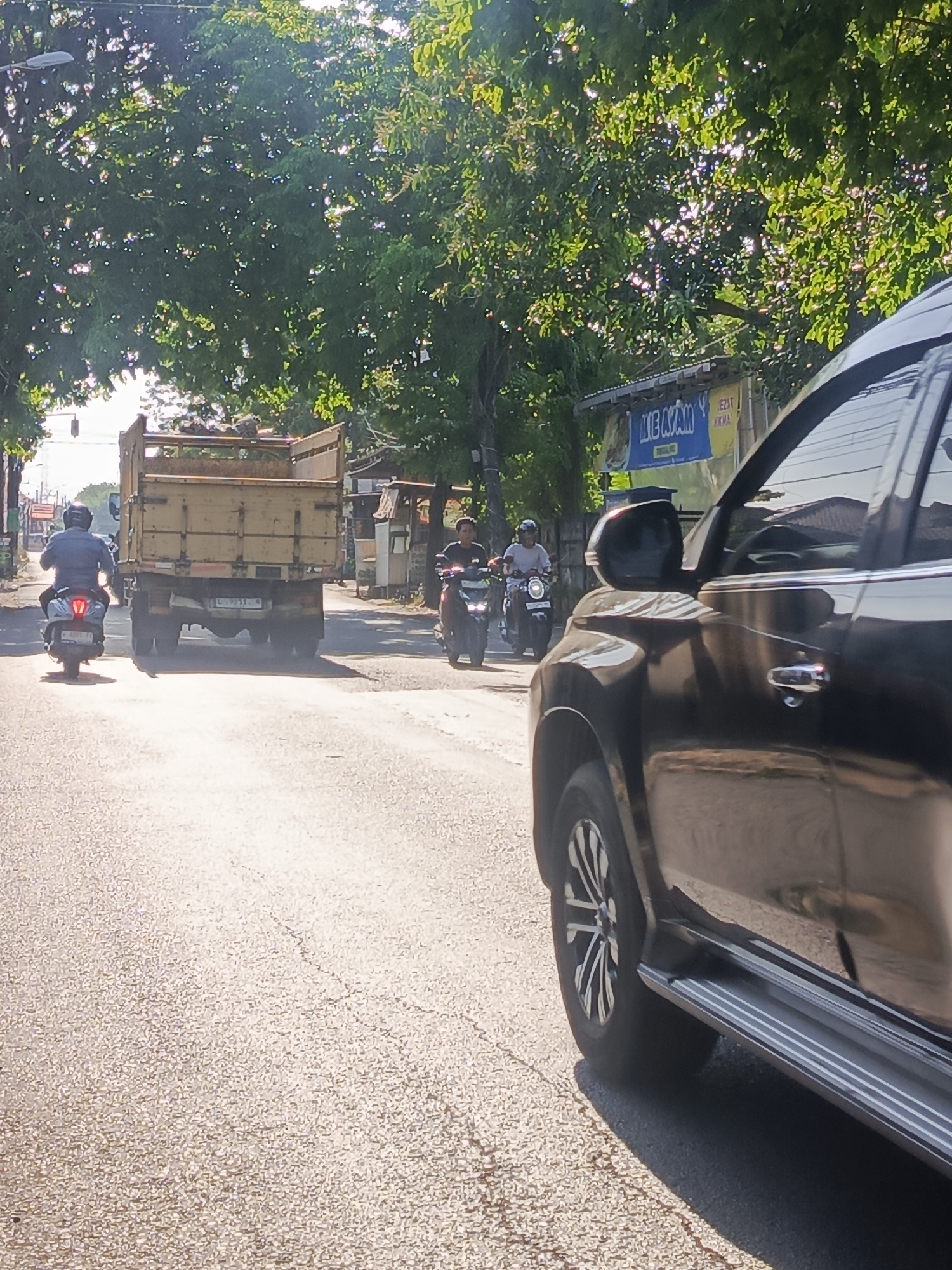 The image depicts a street scene in Setro, Indonesia, likely during daytime given the bright sunlight casting shadows. In the foreground, the right side of a dark-colored SUV is visible, showing its side door, a silver step rail, and a portion of its wheel. The road surface appears to be asphalt with some cracks.
Further down the street, a light-colored truck is traveling. To the left of the truck, a person is riding a scooter, facing away from the camera. To the right of the truck, another scooter is visible with a rider. In the background, lush green trees line the street, and sunlight filters through their leaves. A blue sign with yellow lettering that appears to read "MIE AYAM" (a type of noodle soup) is also visible on the right side, suggesting the presence of a food stall or restaurant. The overall impression is a typical street scene in a town or city in Indonesia, with vehicles and pedestrians on the road.