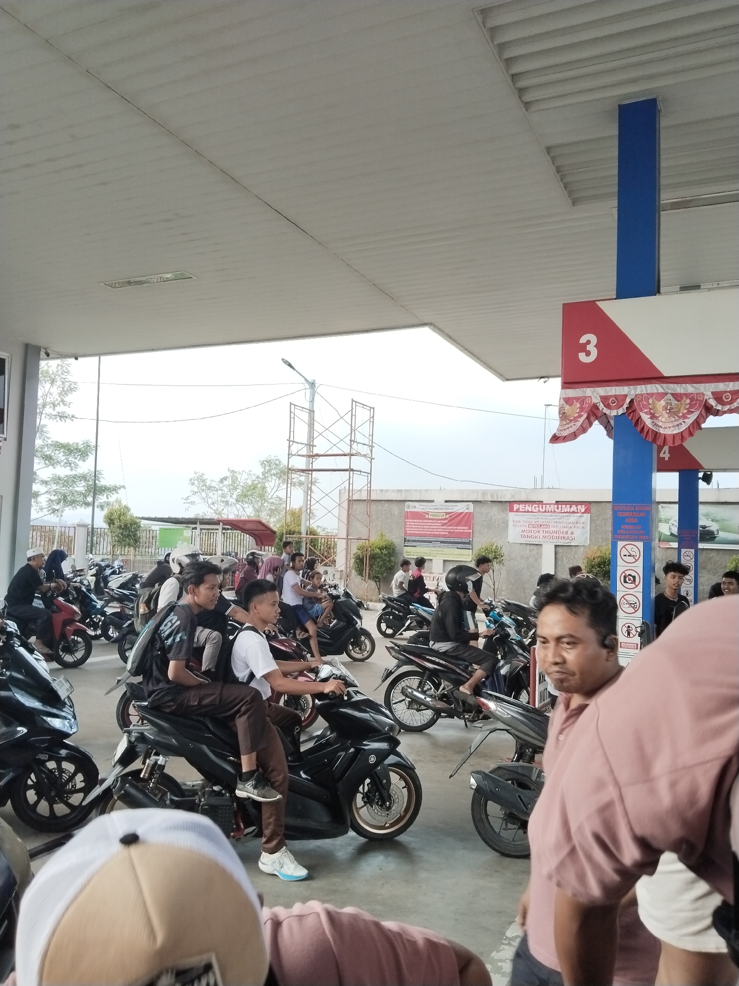 A group of people, mostly young men, are gathered at a gas station with their motorcycles. The gas station appears to be in Sukabares, Indonesia, as indicated by the signage. Many of the individuals are seated on their motorcycles, some are standing, and there is a general sense of waiting or congregating. The environment is an open-air structure with a white, corrugated ceiling supported by blue and red pillars. Various motorcycles are parked in rows, some of which are being ridden or about to be ridden. Banners and signs are visible in the background, including one with the number "3" on it, suggesting multiple service points. The ground is paved, and the overall lighting suggests it is daytime. Some individuals are wearing casual clothing, including t-shirts, long pants, and headwear like caps and hijabs. The focus of the image is on the collection of people and their motorcycles within the gas station environment.