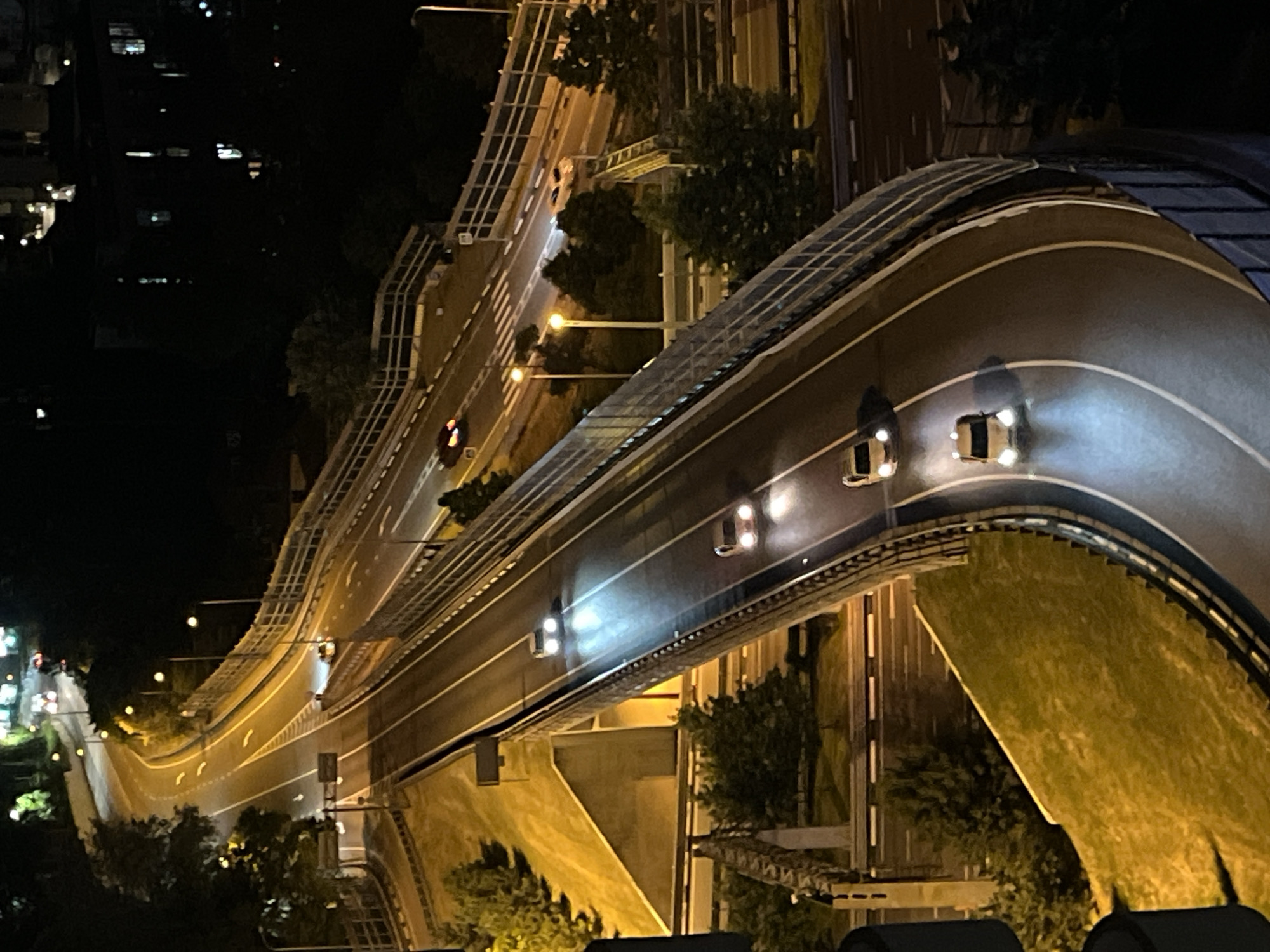 The image is a nighttime aerial view of a multi-level highway or overpass system in Sochi, Russia. Several cars are visible on the illuminated roadways, their headlights creating bright beams. The scene is characterized by the artificial lighting of the road, streetlights, and distant buildings. The environment appears to be urban, with the highway curving through the landscape. There are no people clearly visible in the image, only vehicles in motion. The overall atmosphere is dark, with the lights of the city and vehicles providing the primary illumination.