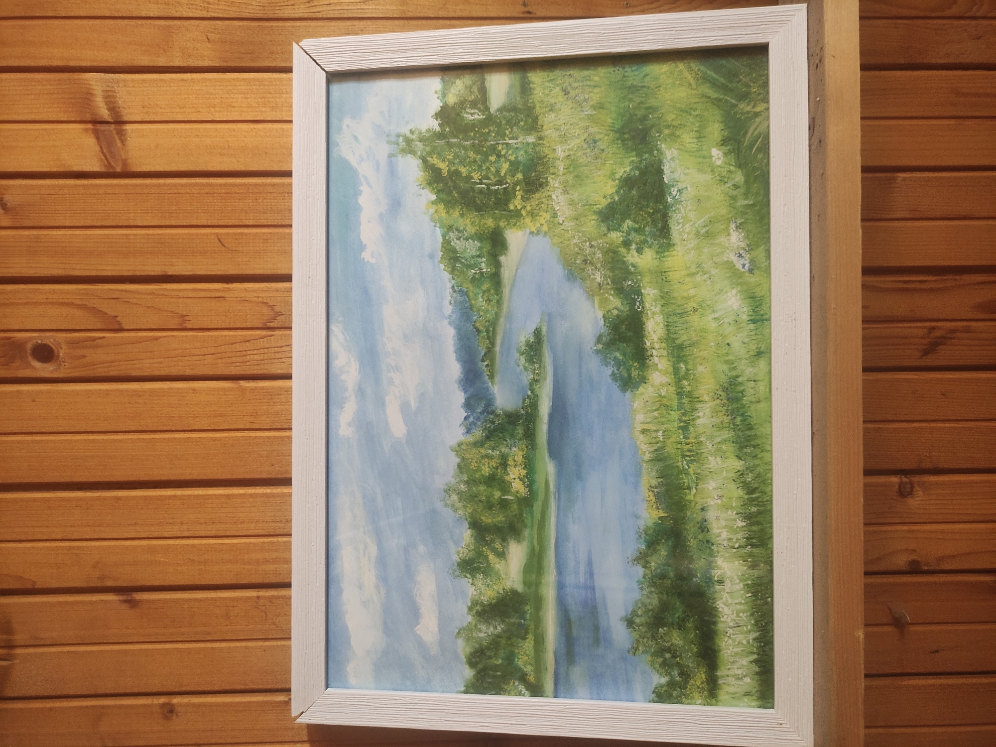 A framed painting is hanging on a wooden wall. The painting depicts a river scene with trees and greenery on either side of the river, with blue sky and white clouds visible. The frame is white with a grooved design. The wooden wall is made of horizontal planks with a reddish-brown tone.