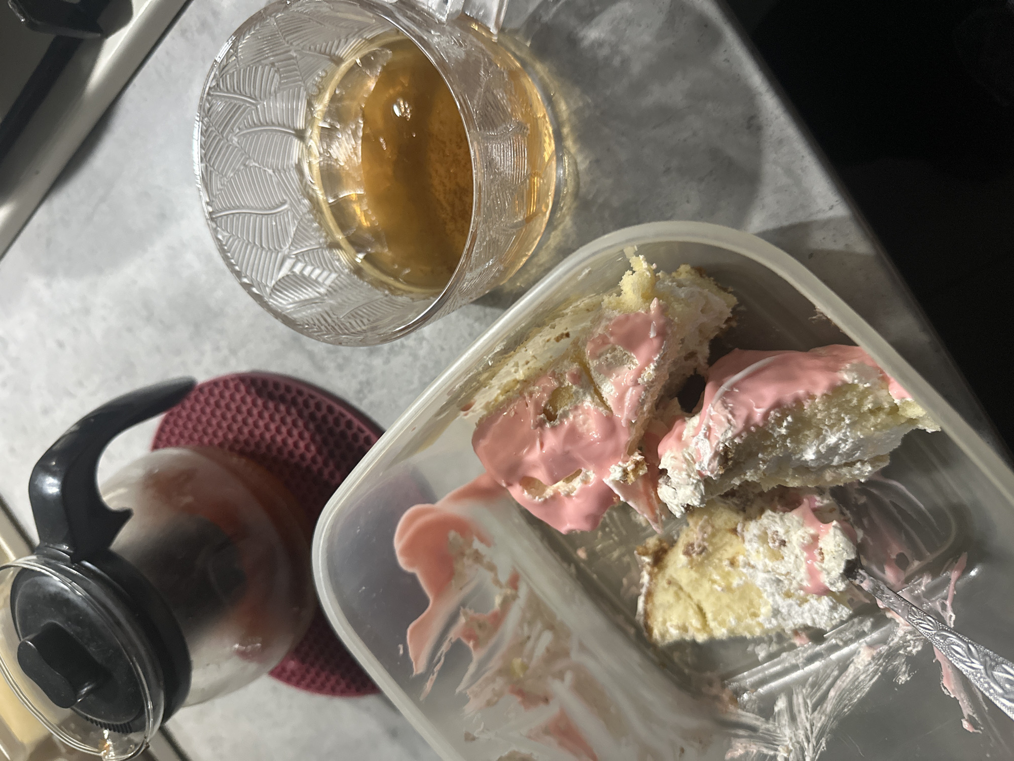 The image shows a close-up view of food and drink items on a light-colored surface, possibly a countertop, in Unknown, Unknown. A partially consumed cake with pink frosting sits in a transparent plastic container, with a silver-colored spoon resting inside. Next to it is a clear glass mug filled with a brownish liquid, likely tea or coffee. A glass coffee pot rests on a red, textured mat. The scene suggests a casual, informal setting, possibly a kitchen or dining area.