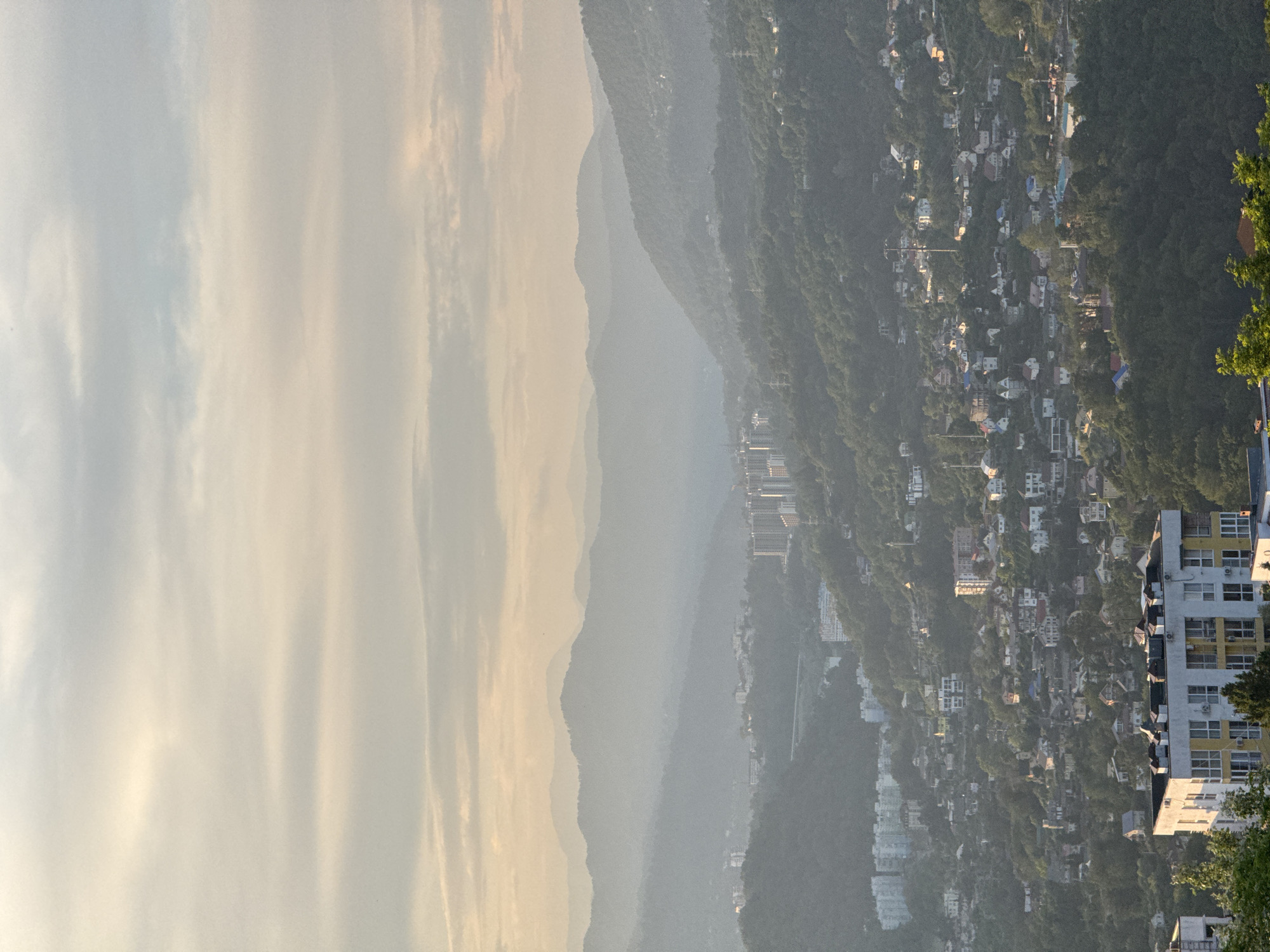 The image depicts a panoramic view of a densely populated hillside settlement in Sochi, Russia, during a hazy twilight. The foreground features a modern, multi-story building with a white facade and yellow accents, overlooking a sprawling landscape of smaller houses and apartment complexes nestled into the verdant, tree-covered slopes. In the mid-ground, several taller apartment buildings and some structures with visible lighting are scattered amongst the residential areas. The background is dominated by multiple layers of hazy, silhouetted mountains that recede into the distance, suggesting a location in the foothills of the Caucasus Mountains. The sky is overcast with soft, diffused light, indicating either sunrise or sunset. No people are visible in the image. The overall impression is one of urban development encroaching on a natural, mountainous environment.