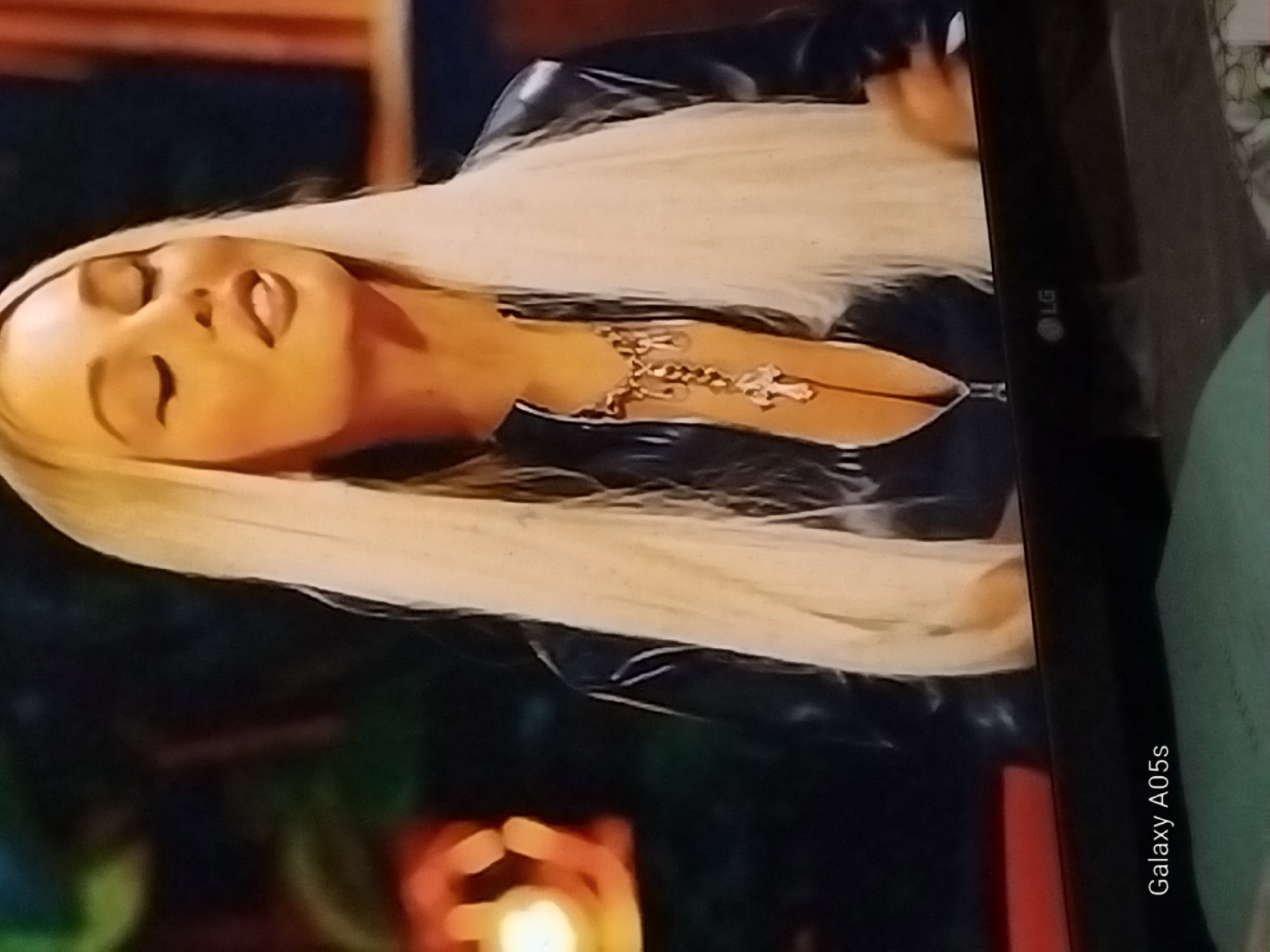 A medium close-up shot shows a blonde woman with her eyes closed, her head tilted back slightly, and her mouth open as if speaking or singing. She is wearing a black, shiny, zippered garment and a necklace with a large cross pendant. Her long blonde hair cascades down her shoulders. The background is dark and blurry, with some out-of-focus orange and yellow lights on the left side. The bottom of the image shows a black surface with an LG logo and the text "Galaxy A05s". The scene appears to be indoors, possibly a television broadcast or a staged performance. There is no indication of the city of Druzhba, Russia.