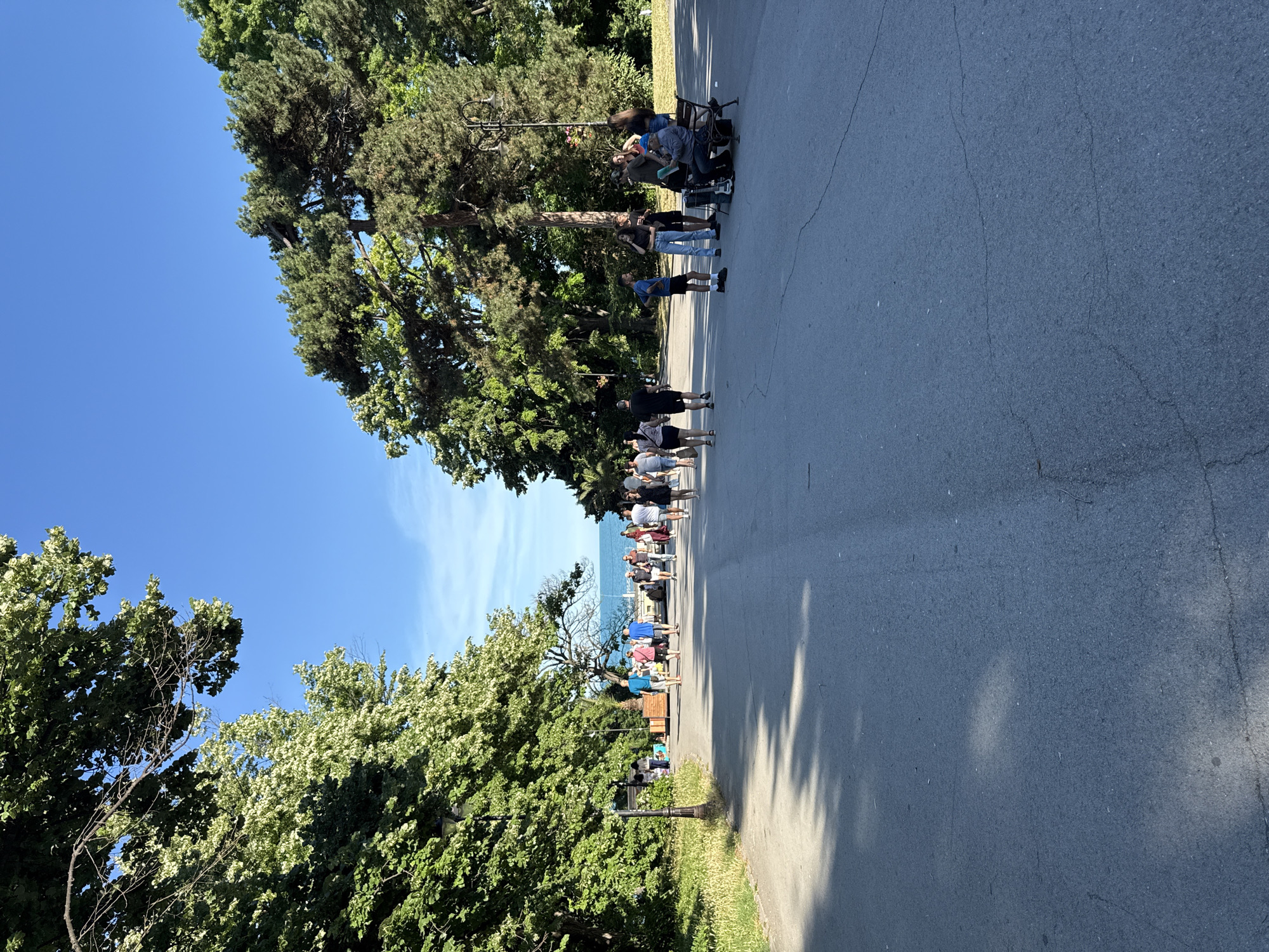 In an outdoor park setting, a paved pathway leads toward a body of water under a clear blue sky. Lush green trees line both sides of the pathway, casting shadows on the ground. Several individuals are walking along the path in both directions, appearing to be enjoying a leisurely stroll. On the right side of the pathway, a group of people are gathered, some seated on a bench, while others stand nearby. One individual on the bench appears to be playing a musical instrument, possibly a guitar, accompanied by another person. The general atmosphere suggests a relaxed and pleasant day in a public park.