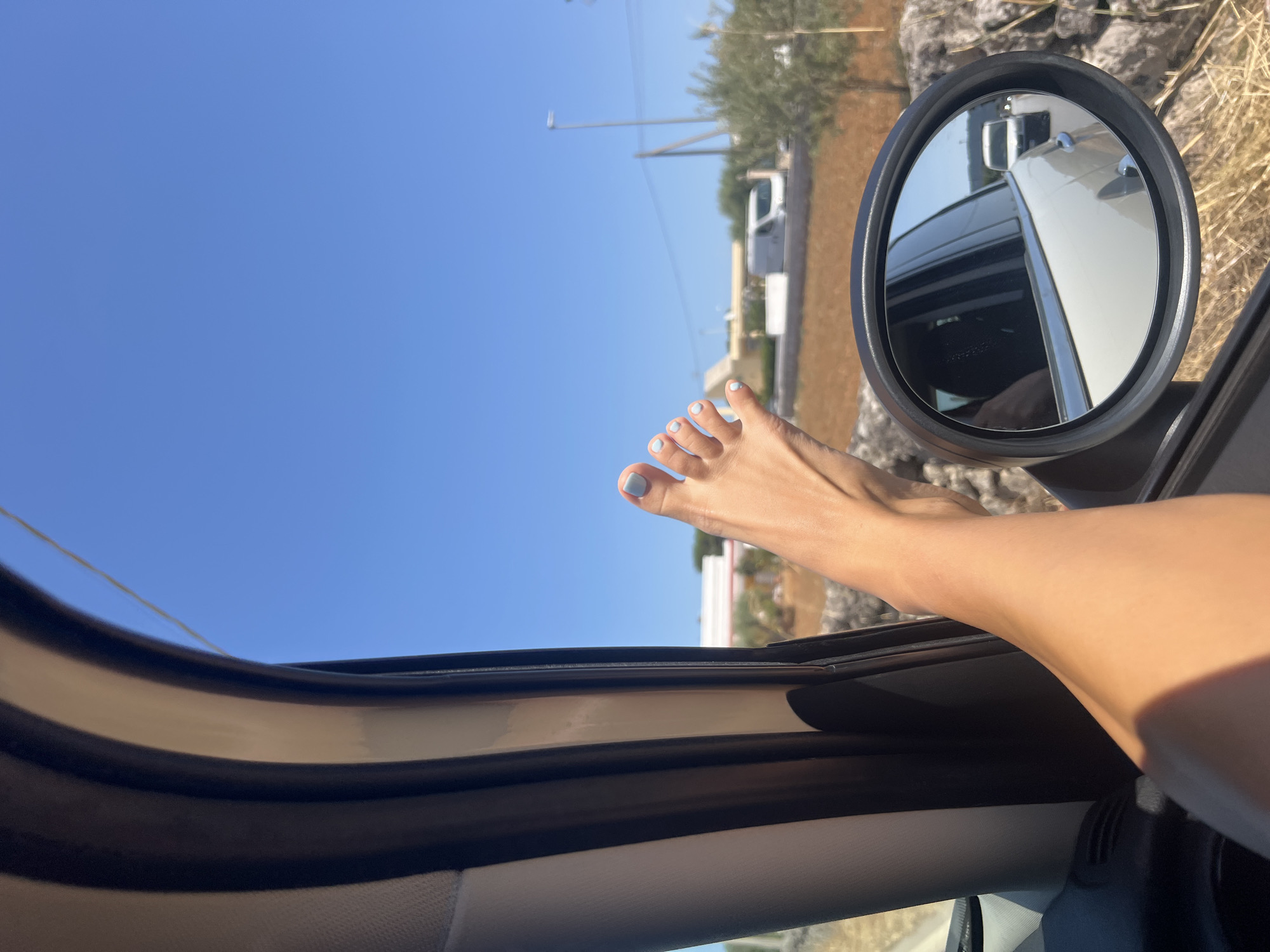 A close-up, eye-level shot shows a person's bare foot with light blue painted toenails resting on the exterior of an open car window. The foot extends out of the frame from the bottom left. The car's interior, including dark and light trim and a side mirror, is visible. The side mirror reflects the road behind, showing a white van and another vehicle. The background is a clear blue sky with a hint of a dry, rural landscape featuring a white building, a street lamp, olive trees, and a stone wall. The scene is likely in Fasano, Italy, given the rural, Mediterranean context.