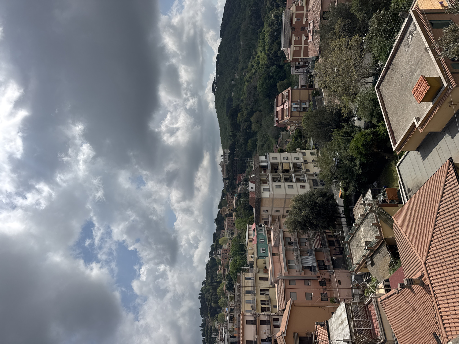 The image presents an expansive, elevated view of a residential town nestled on a hillside, consistent with the location of Ariccia, Italy. The perspective looks across and down a densely built area, with a prominent, heavily forested mountain or large hill rising steeply in the background on the right.
The scene is populated by numerous buildings of varying sizes and architectural styles, constructed along the undulating slopes. Many structures are multi-story apartment blocks and houses, featuring warm, light-colored facades in shades of pink, yellow, and beige. A mix of roof types is visible, with traditional terracotta tiles being prevalent, especially in the foreground and midground, alongside some flatter, grey roofs. Green trees and other vegetation are scattered throughout the urban fabric, particularly on the lower slopes and between buildings, providing a natural contrast to the built environment.
The sky occupies the upper portion of the frame and is largely overcast with extensive, dense grey clouds. Despite the heavy cloud cover, several patches of bright blue sky are visible through breaks in the clouds, indicating a partially cloudy daytime.
No specific activity or event is taking place; the image captures a static, panoramic landscape of the town. The time of day is clearly daytime, illuminated by natural light, but the overcast conditions make it difficult to pinpoint the exact time. No people, animals, or vehicles are visibly engaged in any activity. There is no discernible text on any of the buildings, signs, or objects within the frame. The overall impression is one of an established hillside community under a cloudy sky.