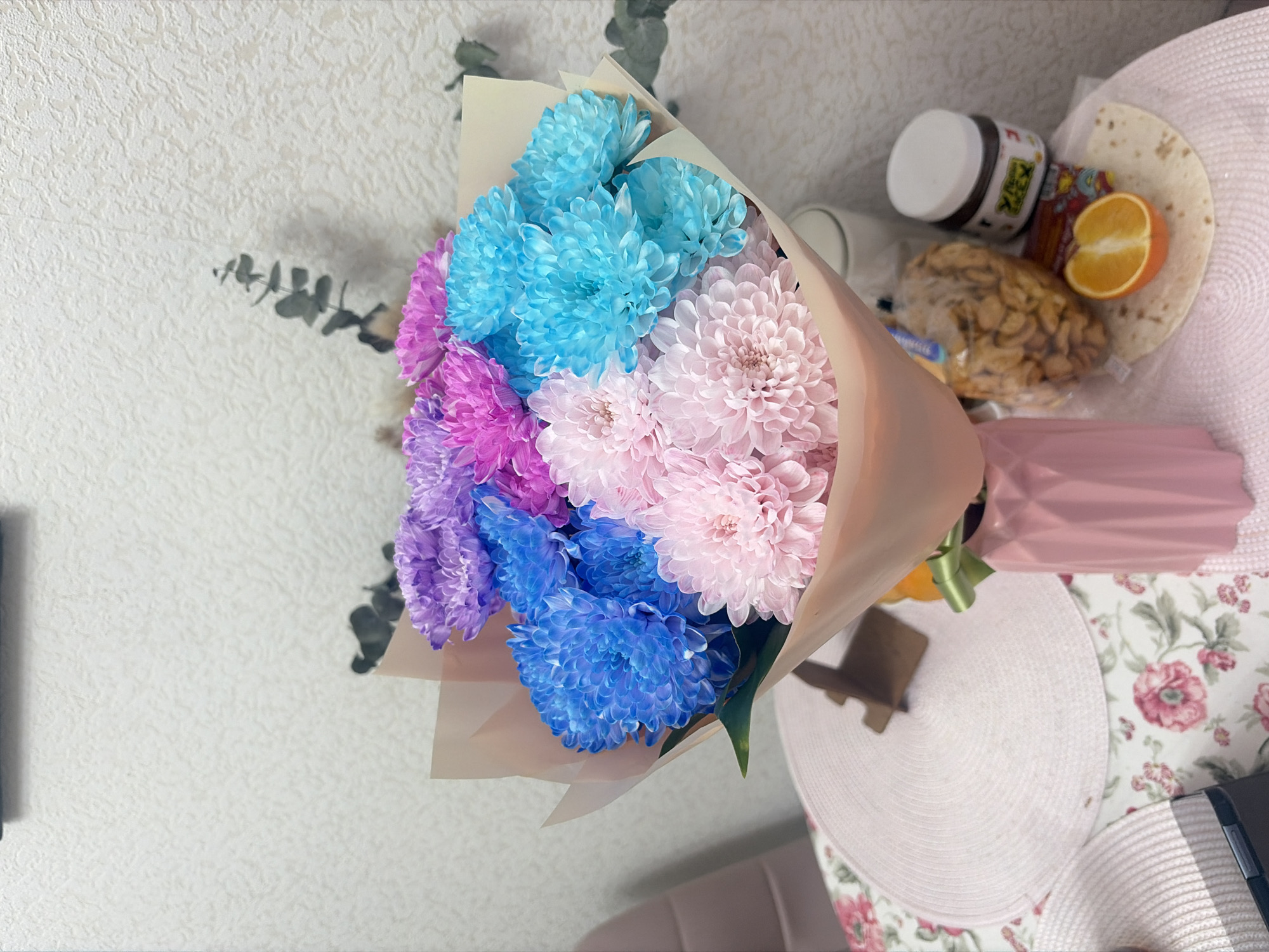 The image depicts a bouquet of pink, blue, and purple chrysanthemums wrapped in tan paper. Next to the bouquet is a collection of items including a white jar of spread, a bag of cereal or trail mix, a halved orange, and what appears to be a tortilla, all resting on a light-colored floral patterned surface with a pink hat. The location of the photograph is not discernible from the provided image. The city of Saint Petersburg, Russia cannot be identified from the provided image. There are no people visible in the image.