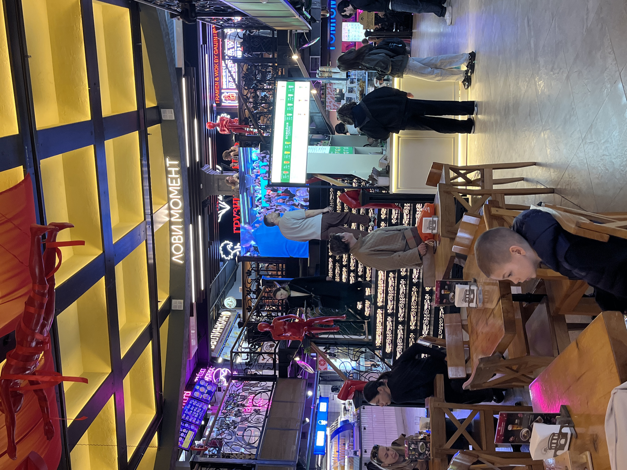 The image is an interior shot of what appears to be a multilevel food court or dining area, probably located in a shopping mall in Moscow, Russia.
The setting is characterized by a mix of seating areas, with people seated at wooden tables and chairs, suggesting they are eating or socializing. Several individuals are visible: a young boy looking down at something, a person in a black jacket, and other customers at a food counter.
The environment is brightly lit with diverse lighting fixtures, including neon signs and ceiling lights. Various food outlets can be identified, with the signs in both Russian and English. In terms of aesthetics, the scene has a modern, eclectic design with architectural elements and art installations.