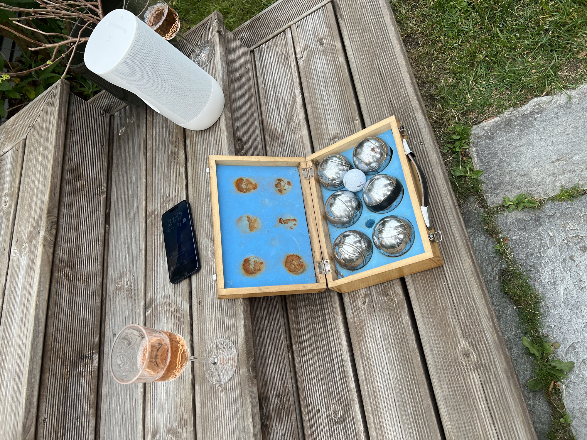 On a wooden deck in Larkollen, Norway, a set of bocce balls lies open in a wooden box lined with blue felt. There are seven metallic balls and one small white ball visible in the box. A glass of rosé wine sits to the left of the box, and a smartphone is placed on the deck to the right of the wine glass. A white, cylindrical speaker is positioned on a higher step of the deck, to the right of the smartphone and also to the right of a second glass of rosé wine. The deck is made of weathered wooden planks. In the background, green grass and foliage are visible, suggesting an outdoor setting.