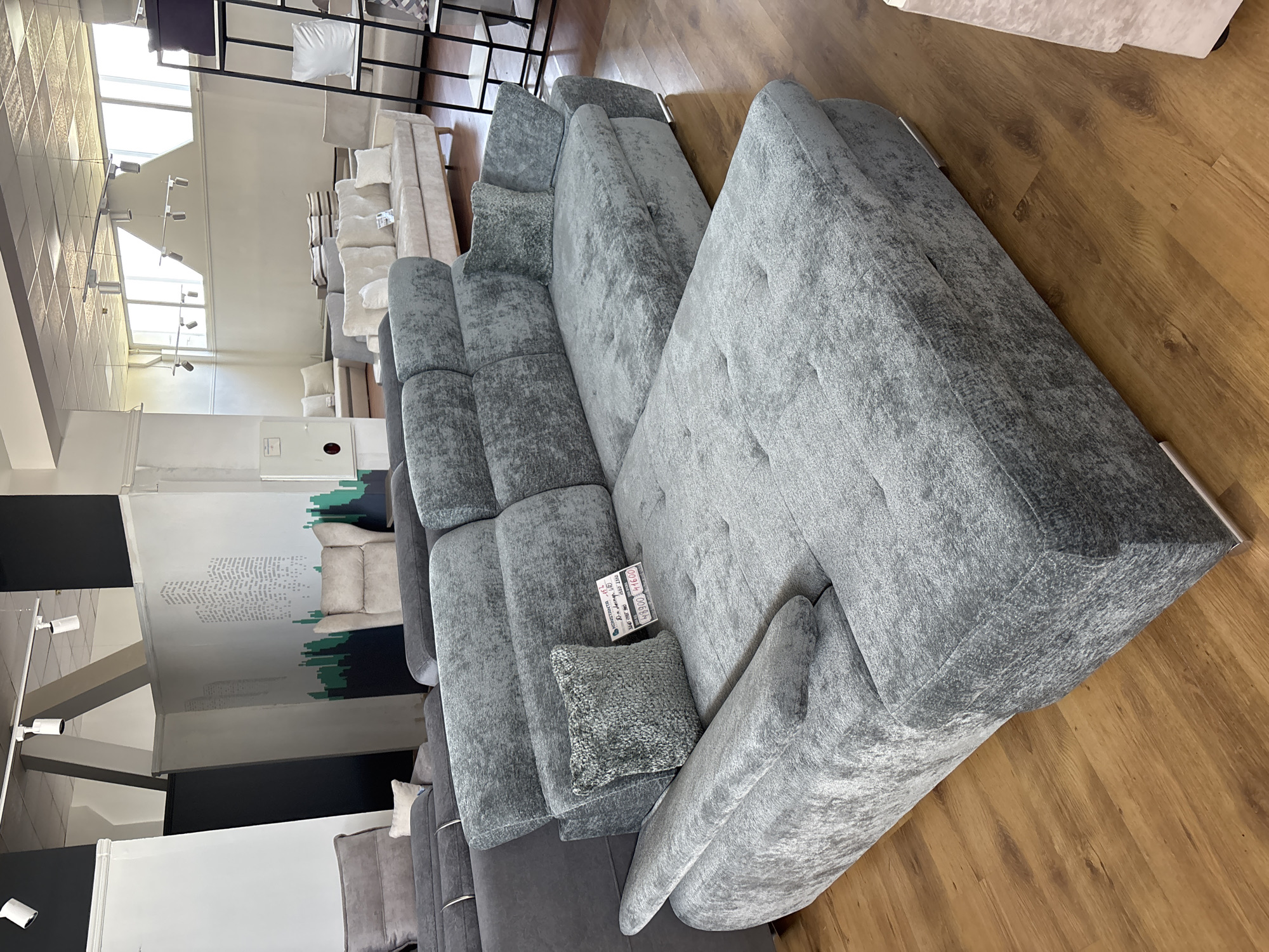 This image shows a large, grey sectional sofa in a furniture store. The sofa is upholstered in a textured fabric and has several cushions. It is arranged in a modular configuration, with multiple seating sections and a chaise lounge. A small, decorative pillow sits on one of the sections. A price tag is visible on the sofa, but the text is too small to read clearly. The sofa is placed on a wooden floor, and in the background, other furniture and shelving units are visible. The setting appears to be a well-lit showroom, possibly in Zaporizhzhia, Ukraine, given the provided location context. There are no people visible in the image. The overall scene depicts a retail environment showcasing furniture for sale.