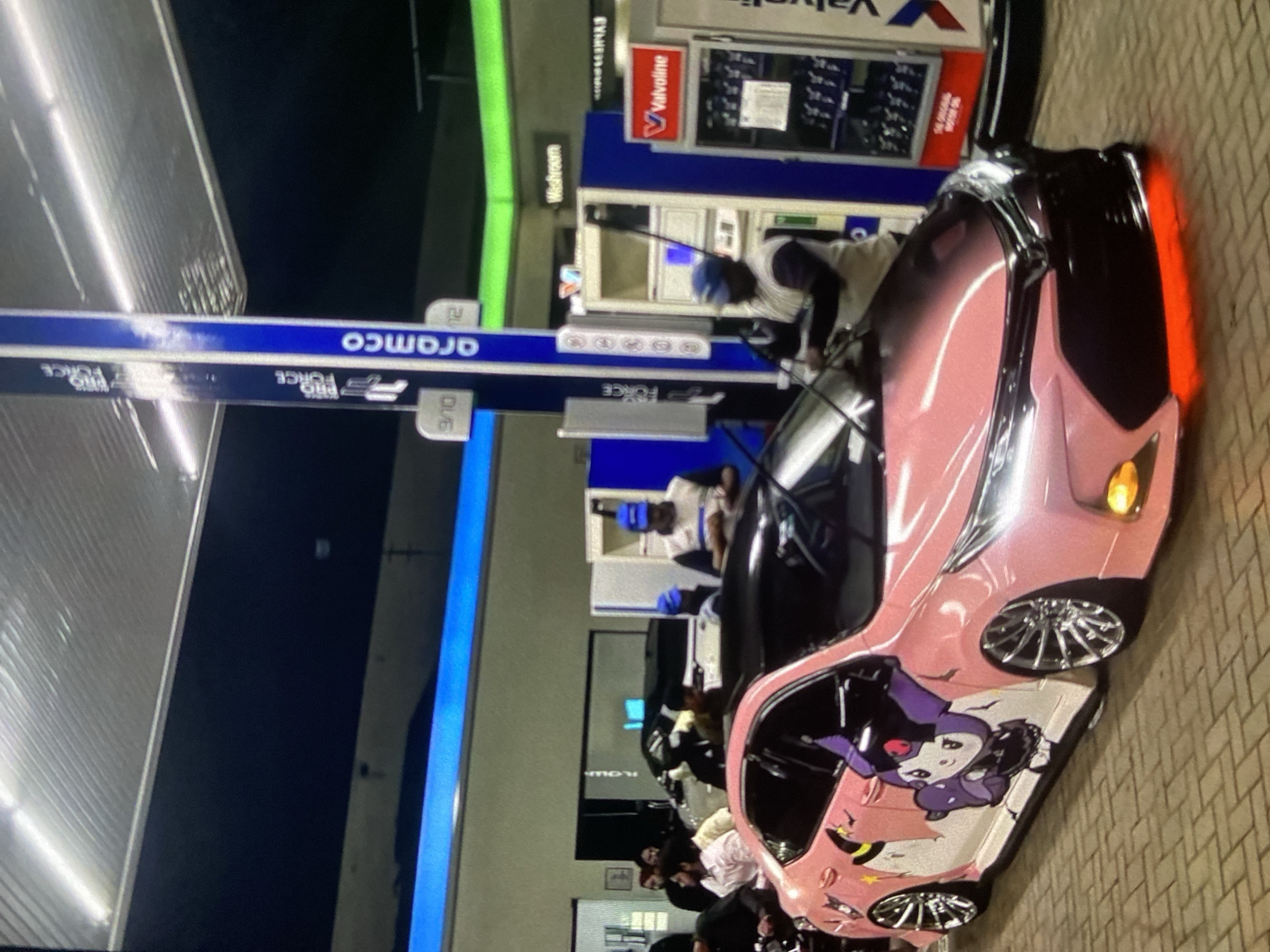 The image depicts a pink car decorated with anime characters at a gas station. Aramco and Valvoline signage are visible, and people are gathered around the car. The image is rotated, and the location details of Lhokseumawe, Indonesia, are not evident in the photograph.