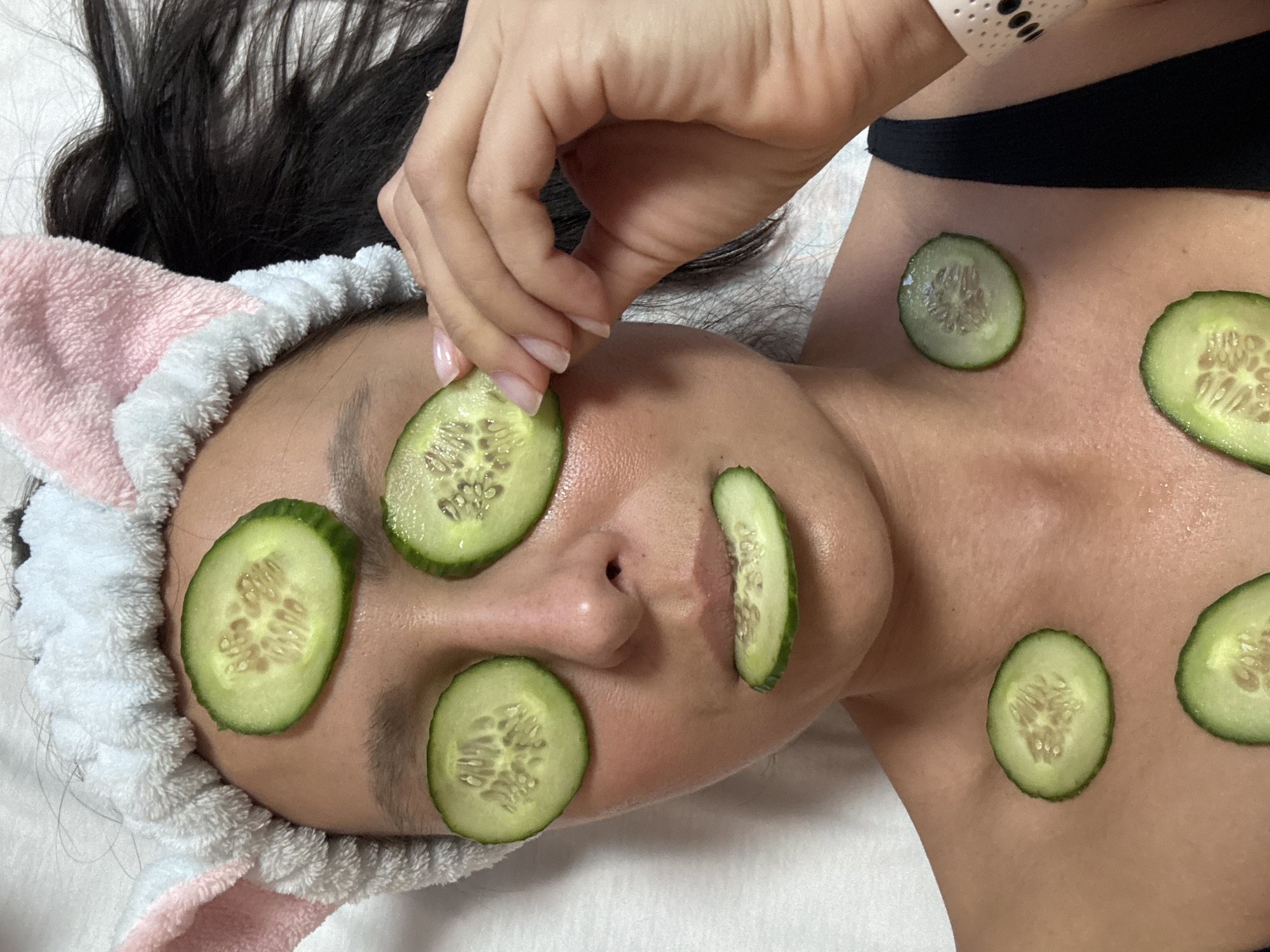 An overhead shot captures a person, likely a woman, undergoing a relaxation or beauty treatment in Chaikovsky, Russia. The individual is lying on a white surface, possibly a spa bed or clean sheet.
Multiple fresh cucumber slices are placed strategically on the person's face and upper chest. Two slices cover the eyes, another rests on the forehead, one on the cheek, and three more are visible on the décolletage area. The person's right hand, with neatly manicured nails featuring a light pink polish, is shown in the process of placing or adjusting a cucumber slice on the forehead.
The person wears a distinctive fluffy headband, pink and white with what appear to be bunny ears, holding back their dark hair. A black strap, possibly from a top or bra, is visible near the right shoulder. The skin on the face and chest has a slight sheen, suggesting moisture or a product application. The scene evokes a sense of tranquility and self-care. No text is visible in the image.