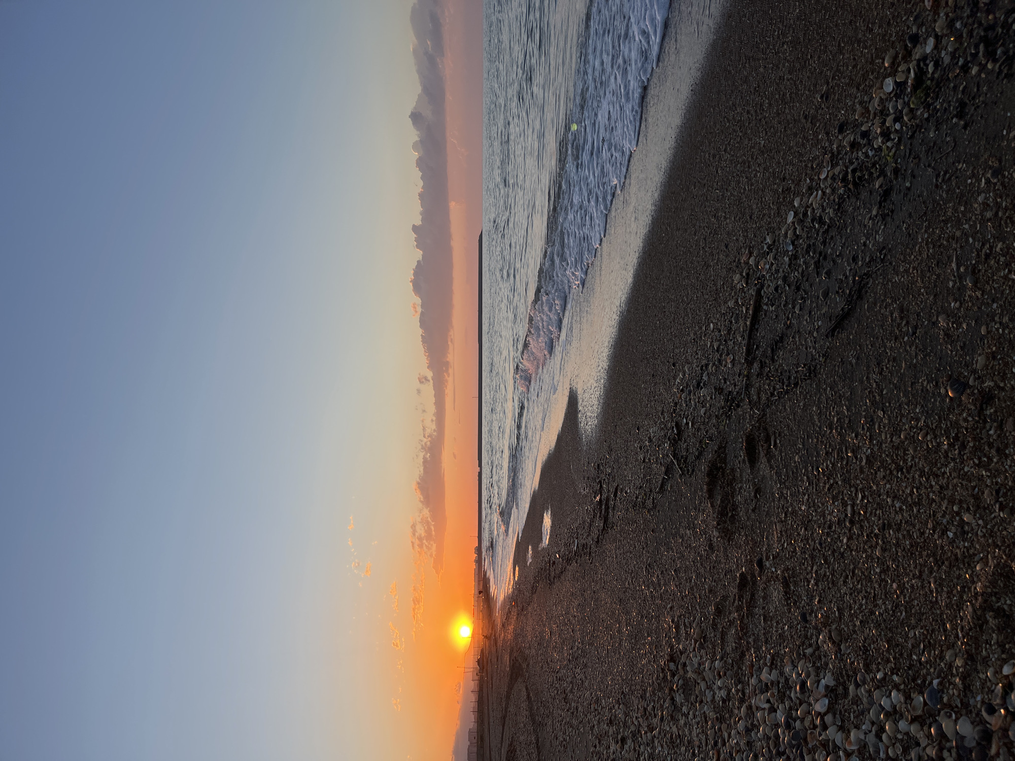 The image shows a beach at sunset in Kaspiysk, Russia. The sun is low on the horizon, casting an orange glow on the sky and water. The beach is made of dark sand and pebbles. Gentle waves are lapping the shore. A few clouds are visible in the sky. The scene is still and tranquil. There are some buildings and a fence to the left of the sun. Footprints are visible in the sand.