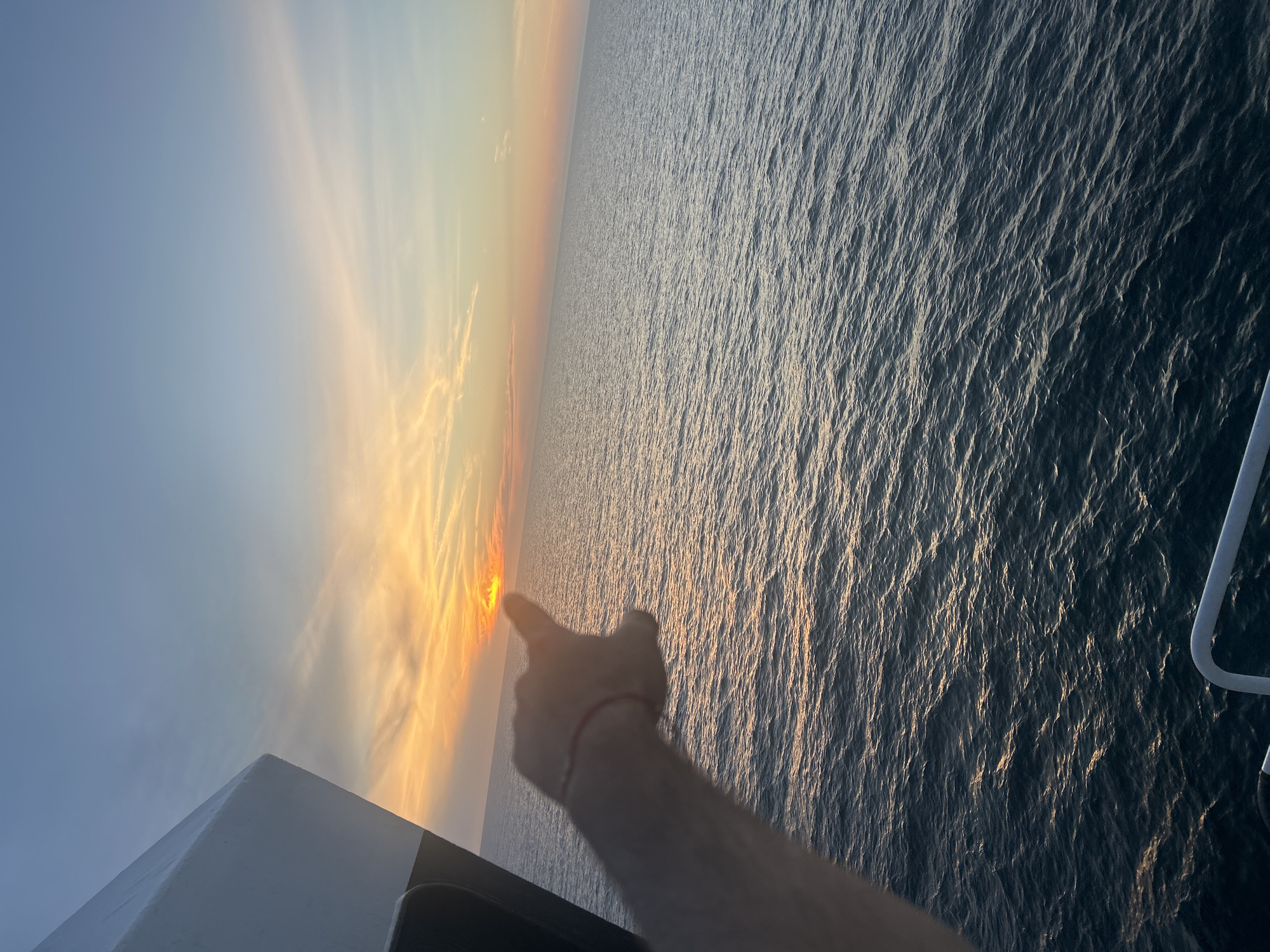 An over-the-shoulder shot shows a person's hand, adorned with a red string bracelet on the wrist, pointing towards the horizon. The hand is positioned in the foreground, with the finger extended towards a sunset over the ocean. The sun is a bright orange disc just above the water, casting a warm glow across the sky, which transitions from yellow and orange near the horizon to blue at the top. Wispy clouds streak across the sky, catching the sunset's light. The ocean below is calm, with small ripples reflecting the sky's colors. On the left side of the frame, a portion of a white structure, possibly a ship or a building, juts into the scene. The overall environment is an open body of water at sunset.