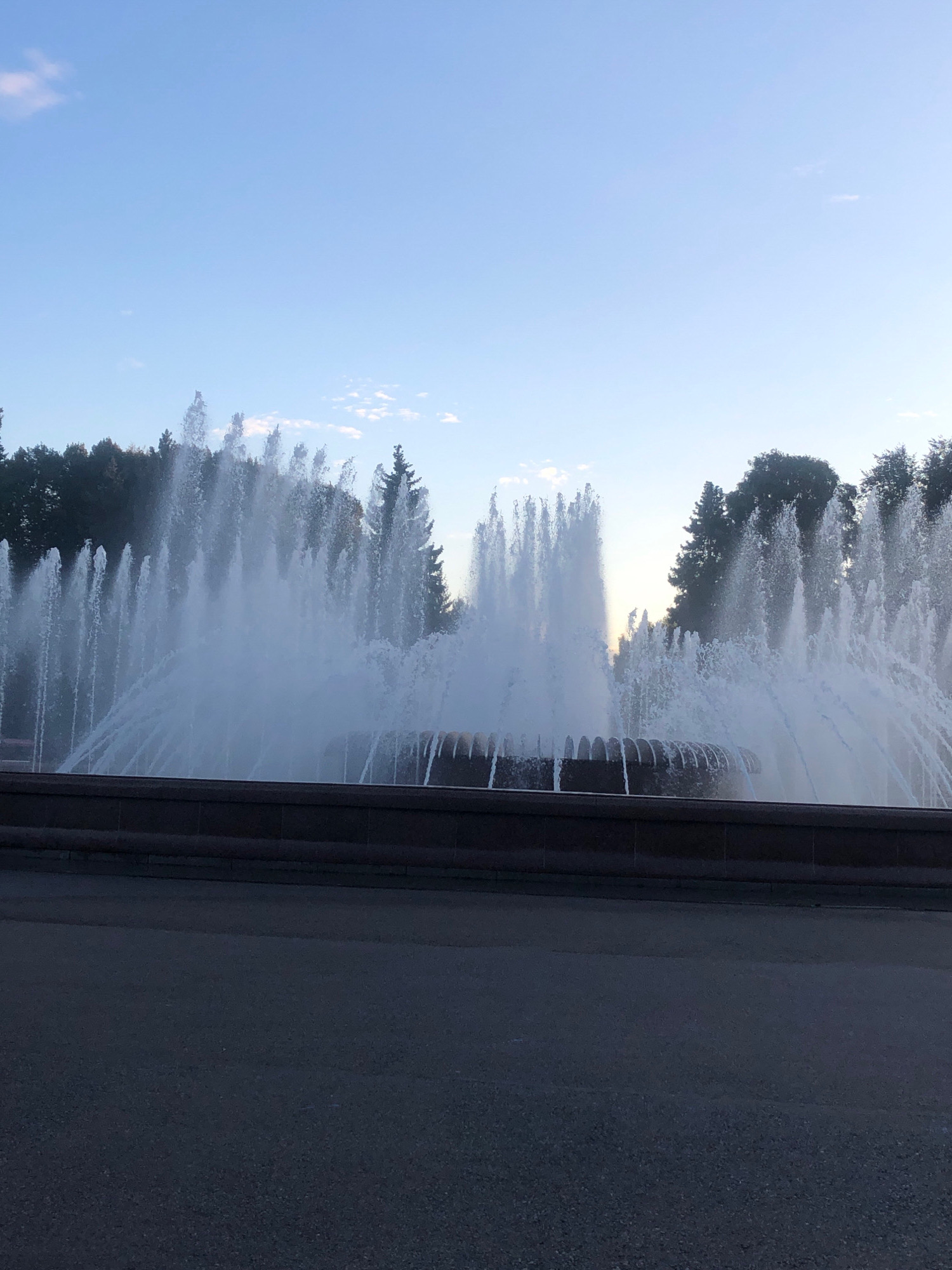 The image depicts a large fountain with multiple jets of water spraying into the air, creating a misty atmosphere. The fountain is set against a backdrop of trees and a clear blue sky with a few wispy clouds. In the foreground, there is a paved surface, likely a walkway or road, and a dark stone border or wall surrounding the fountain. The lighting suggests it is either early morning or late evening. This fountain is located in the park of Victory of Leningrad in the city of Saint Petersburg, Russia. There are no people visible in the image.