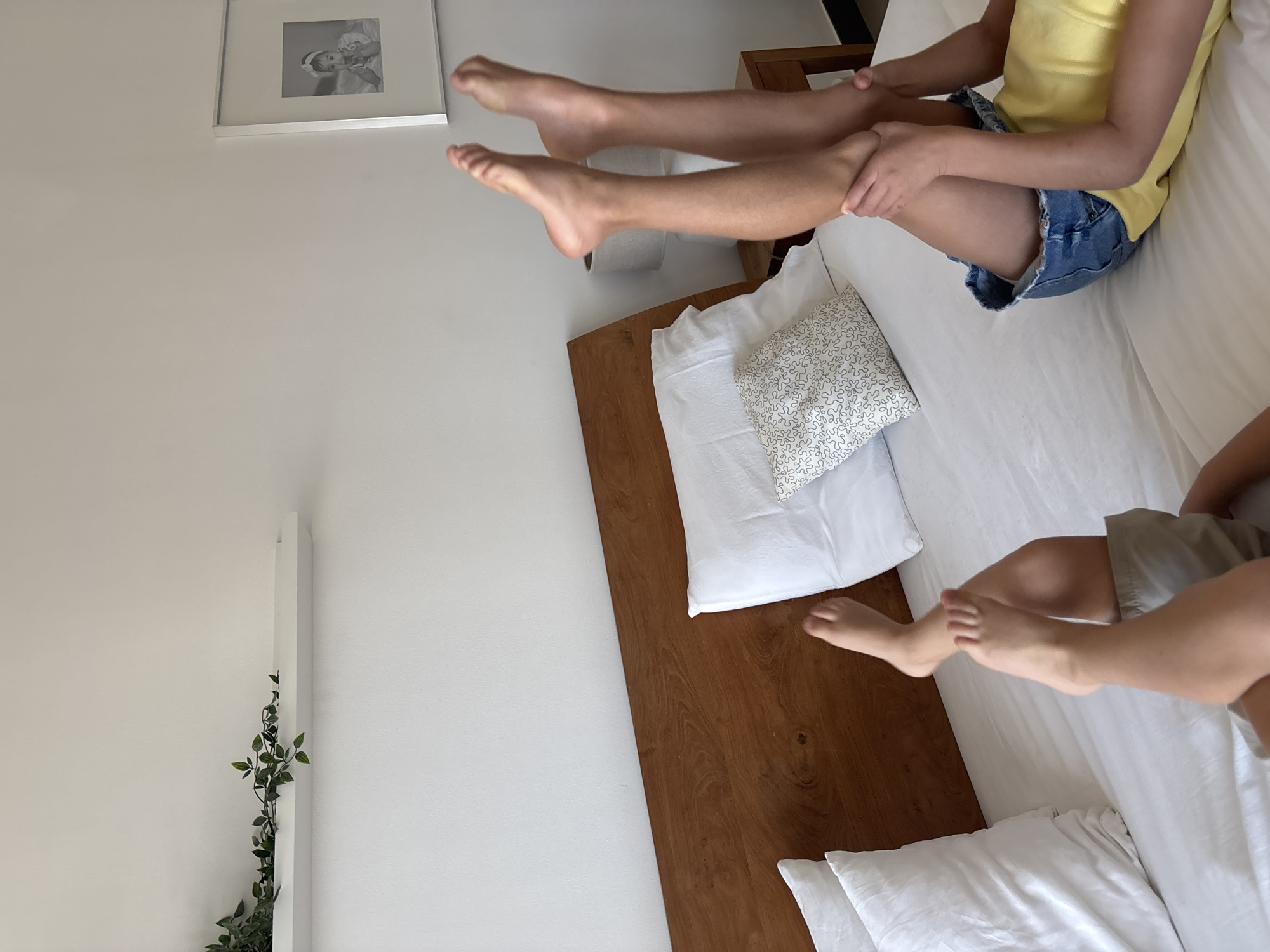 The image shows two people on a bed. One person is wearing a yellow tank top and blue denim shorts, and is sitting on the bed with their legs extended. Their hands are resting on their legs. The other person's legs and feet are visible, extended in the air above the first person's legs. They are wearing grey shorts.
The scene appears to be an indoor setting, possibly a bedroom, with a white wall in the background and a wooden headboard of the bed. There is a framed picture on the wall and some artificial greenery on the left side of the frame. The lighting suggests daytime. The overall impression is one of relaxation or playfulness.
No text is visible in the image. The location context is Dubai, United Arab Emirates.