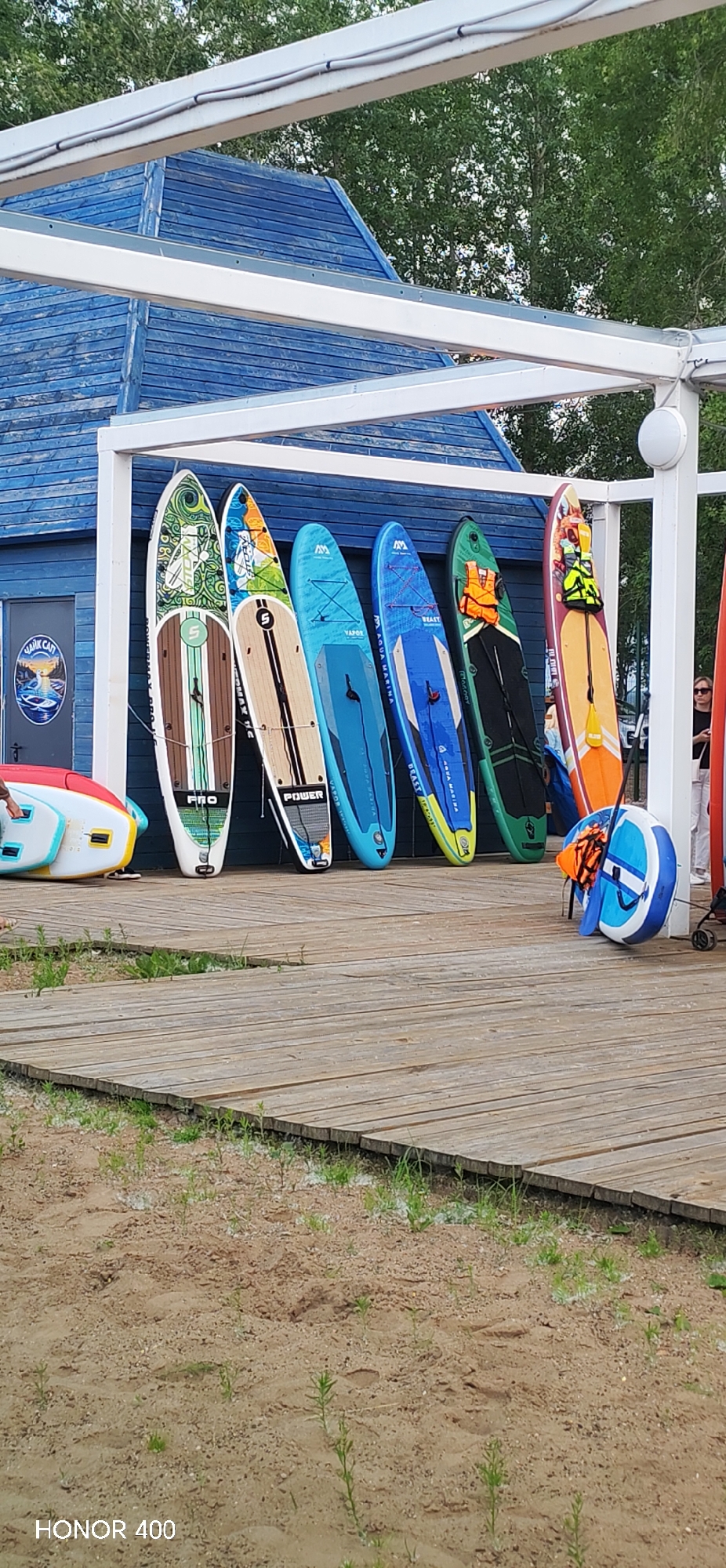 An outdoor scene shows a row of paddleboards leaning against a blue wooden building. The paddleboards are of various colors and designs. Some have graphics, while others have simpler patterns. An orange life vest is attached to one of the paddleboards. In front of the building, there is a wooden walkway that leads to a sandy area with sparse green vegetation. A white and blue inflatable object with an orange life vest attached is also visible on the walkway. The building appears to be a shed or small structure, possibly part of a watersport rental facility. The overall setting suggests a recreational area near a body of water, consistent with the city of Chaikovsky, Russia, known for its proximity to the Kama River and its recreational opportunities. The sky is overcast, and the lighting suggests it is daytime. The image has a watermark at the bottom left corner reading "HONOR 400".