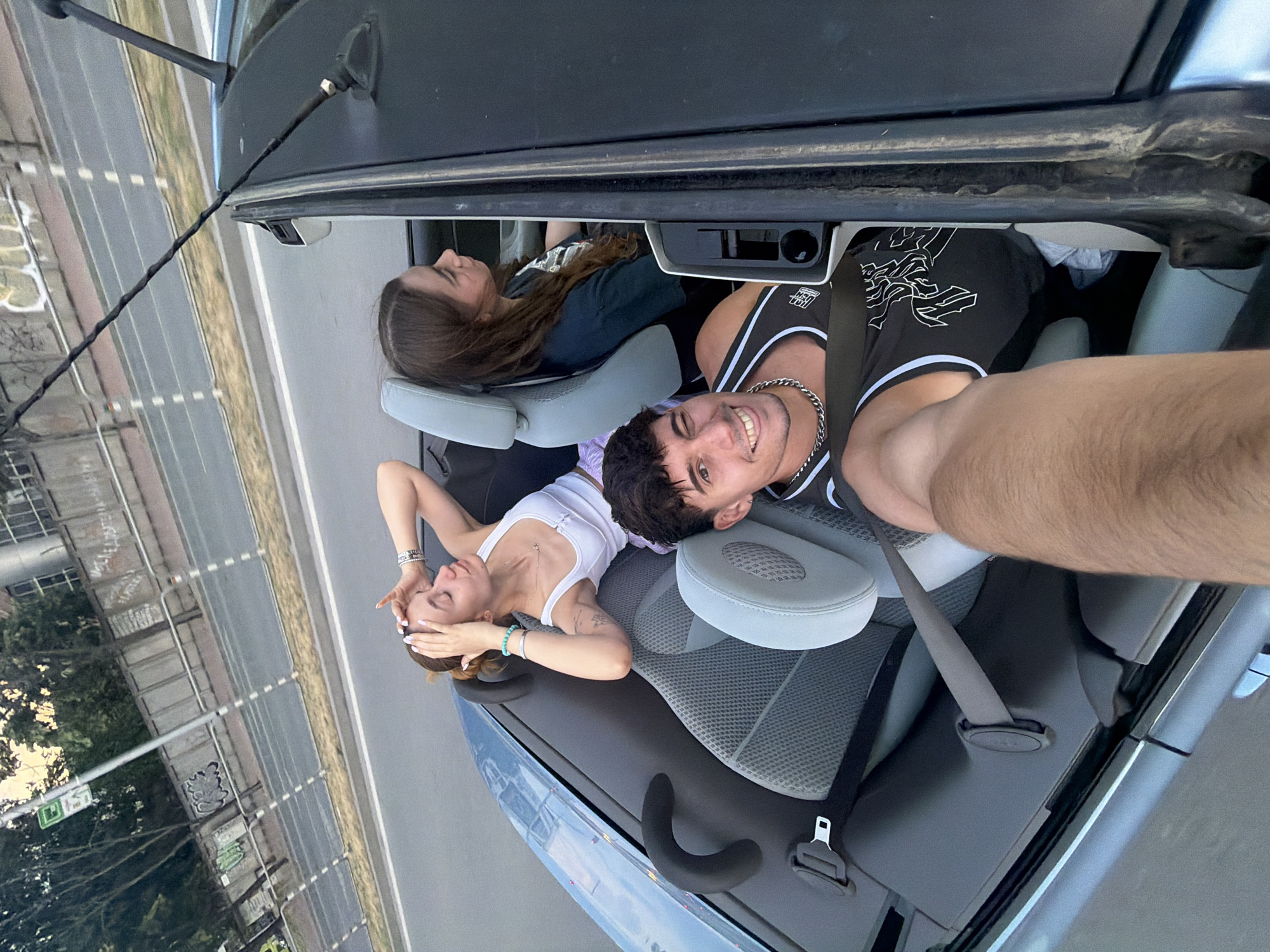 Three individuals are captured in a light blue convertible car in motion on a multi-lane asphalt road in Plovdiv, Bulgaria. The image is taken from a high angle, likely a selfie by the male in the front passenger seat.
The male in the front passenger seat occupies the bottom right foreground, visible from the chest up. He has dark hair, light skin, and is wearing a black tank top with white trim and a white graphic design. He smiles and winks at the camera, holding the camera/phone with his outstretched right arm, which is partially visible. A silver chain necklace is around his neck.
In the rear passenger seat, directly behind the driver's side (left from the viewer's perspective), a female with brown hair and light skin is seen from the shoulders up. She wears a dark blue-green t-shirt and looks forward or slightly to her right, not directly at the camera. Her expression is neutral.
To her right, in the other rear passenger seat, a second female with reddish-brown hair and light skin is visible. She wears a white tank top and light purple shorts. Her hands are positioned on her head, with white nail polish visible. She has silver and blue beaded bracelets on her left wrist. Her eyes are closed, or she is looking down and to her left.
The car's interior features grey fabric seats, light grey headrests, and black plastic trim. A grey seatbelt is buckled on the rear left side, and a black looped handle is on the rear left interior panel. The convertible top is down. A twisted black antenna is attached to the upper right side of the windshield frame, and another antenna-like object is visible on the rear right side of the car.
The background shows an urban environment. The car is on a road bordered by a low concrete barrier, behind which stands a taller concrete wall covered in extensive graffiti, including white, green, and grey tags. Beyond the wall, green trees and bushes are visible. In the upper right background, a large building with rectangular windows is present, possibly an office or industrial structure. A green rectangular sign, likely a road sign or bus stop sign, is visible in the upper left background. The sky is overcast with a soft, warm light, suggesting late afternoon or early evening.
