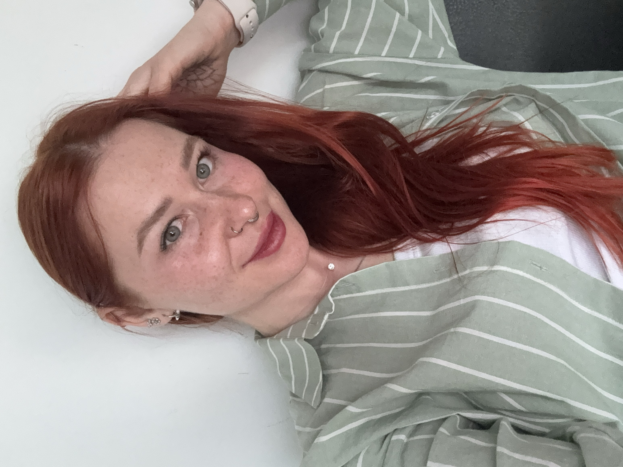 A close-up, slightly angled indoor selfie captures a young woman with long, vibrant red hair spread out around her. She is lying down, looking directly at the camera with a calm expression and a subtle smile. Her fair skin is adorned with scattered freckles, and she has striking green or hazel eyes. Her lips are painted with a dark pink or berry-toned lipstick. Visible facial piercings include a septum ring and a nostril ring. Her left ear features multiple earrings, including a small hoop and a cluster of studs. A small, faded tattoo with an intricate pattern is visible on her upper left arm.
She is wearing a light green collared shirt with thin white vertical stripes over a white top. A delicate necklace with a small, shiny pendant (possibly a diamond) is visible around her neck. A light-colored smartwatch is worn on her left wrist. The background is predominantly a plain white surface, suggesting she is lying on a bed, sofa, or floor, with a darker, indistinct area visible in the upper right corner, likely part of the room's interior. The lighting is bright and even, indicative of an indoor setting during the daytime. No text is visible in the image. The location context provided is Rostov-on-Don, Russia, though no specific visual cues confirm this.