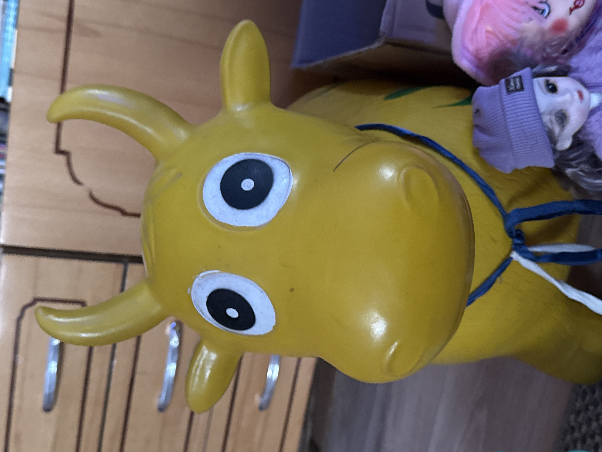 The image features a close-up of a yellow, inflatable, toy horse head, likely a bouncy ride-on toy. The head is prominently displayed, with cartoonish eyes that have black pupils surrounded by white. The horse has short, rounded horns. A blue rope is tied around the horse's neck. Partially visible behind the toy horse are several dolls and a cardboard box. The background includes a light brown cabinet or wall with vertical grooves. The location, Дойная, Russia, is not visually discernible from the image.