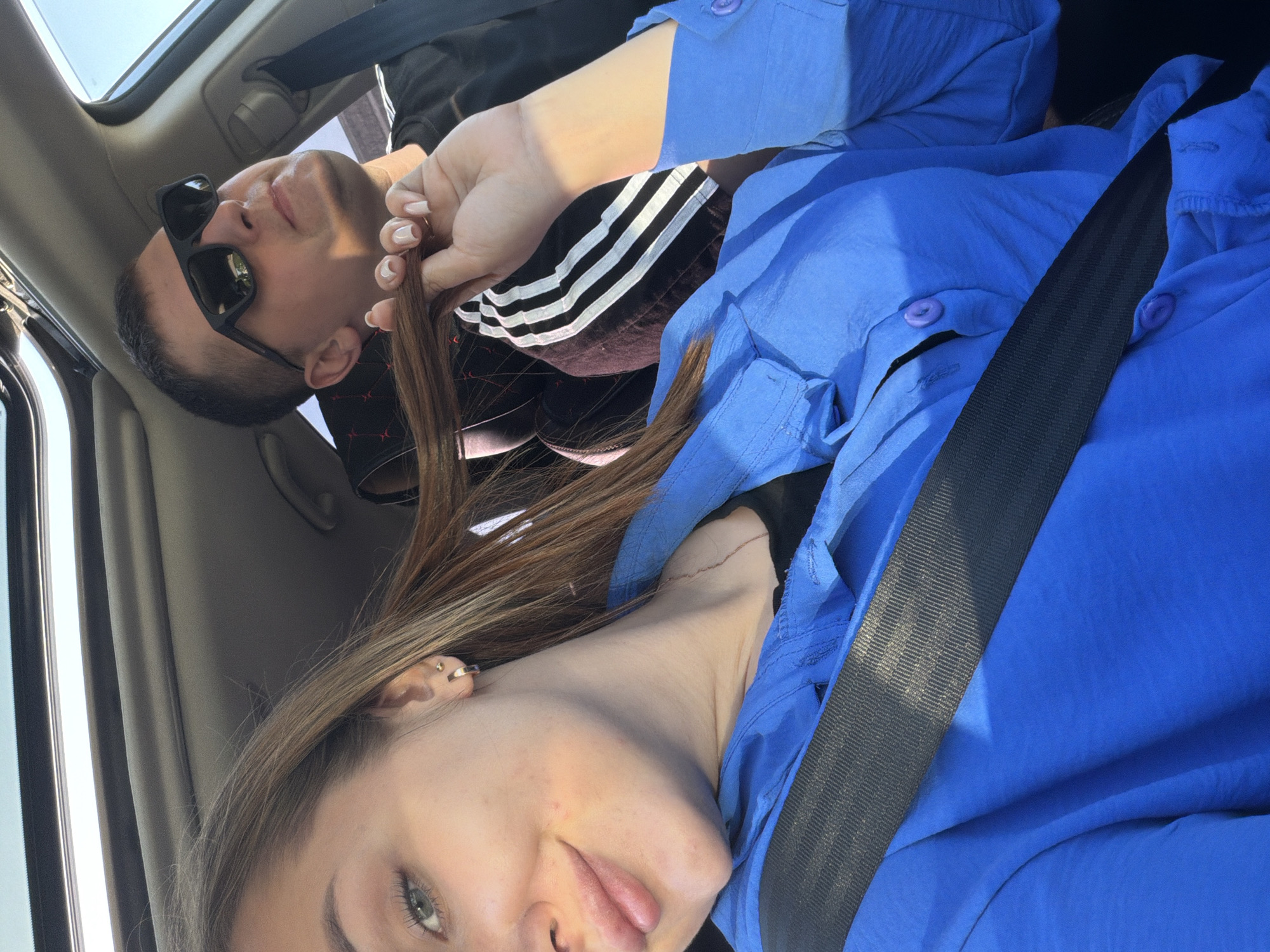 The photograph shows two individuals in what appears to be a car. The image is taken from a tilted perspective. The foreground shows a young woman with light brown hair, wearing a blue button-down shirt and a seatbelt. Her complexion is fair, and she has green eyes and what appears to be a small piercing on her ear. Behind her, there is a man wearing sunglasses and a black Adidas jacket with white stripes. His hair is dark, and he has a medium complexion. He is looking upwards. A strand of the woman’s hair is being held by a hand, likely the woman’s. The car interior is visible, including the ceiling and door. There is no indication of the city of Chaikovsky, Russia in the image. The action appears to be a casual moment in the car.