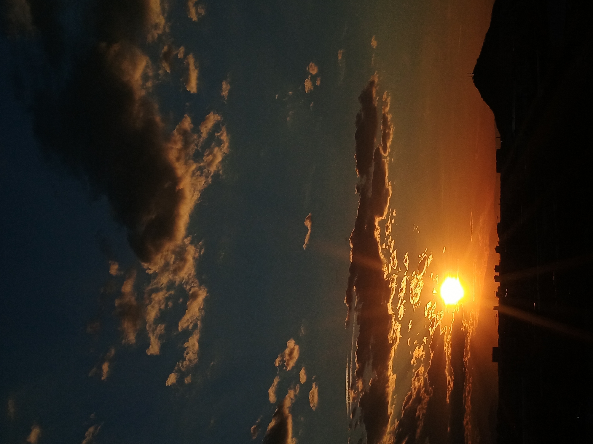 The image depicts a sunset in Plovdiv, Bulgaria. A bright, glowing sun is partially obscured by clouds near the horizon, casting a golden-orange light. Silhouetted buildings are visible on the right side of the frame. The sky ranges from a deep blue on the left to a warm orange towards the sun. Clouds are scattered throughout, some illuminated by the setting sun.