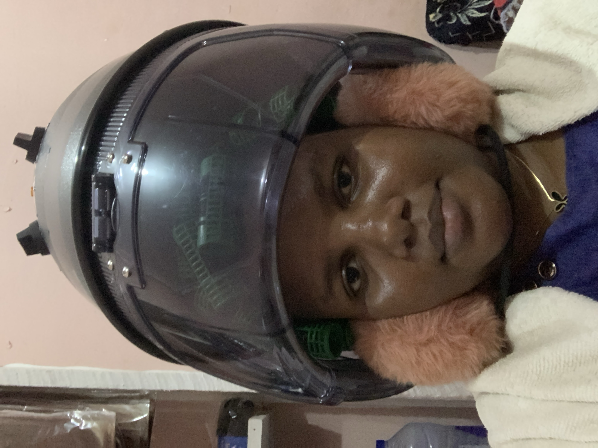 A medium shot shows a woman facing forward, sitting under a hair dryer. The dryer is a domed, dark grey helmet-like apparatus with a clear visor, revealing green hair rollers and peach-colored earmuffs around her face. Her skin has a slightly oily sheen. She wears a blue top and a white towel is draped around her shoulders. A gold necklace is visible at her neck. The background is a peach-colored wall with a white surface visible below, partially covered with wrapped items and bottles. It is not possible to determine the city of Dar es-Salaam, Tanzania from this image alone.
