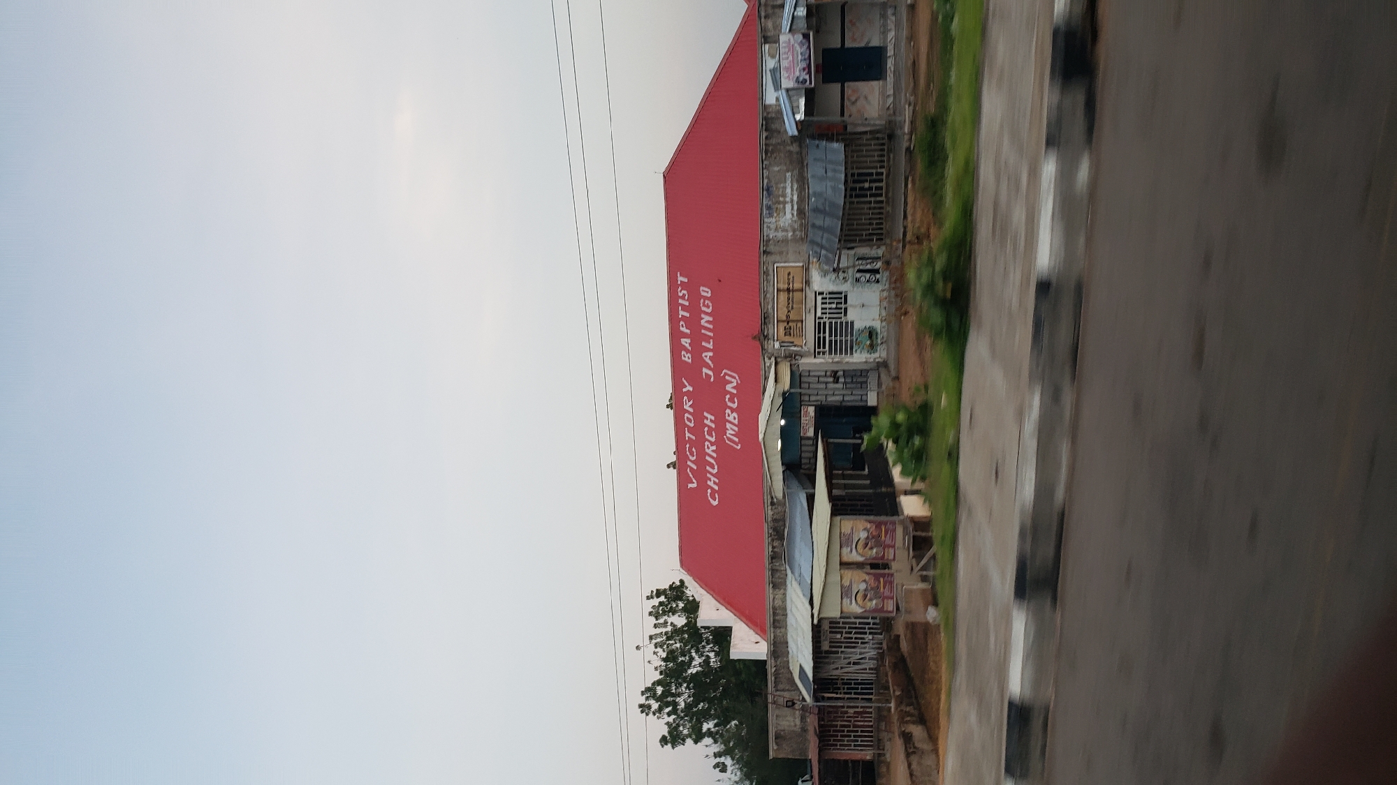 The image shows a side view of the Victory Baptist Church in Jalingo, Nigeria, with a red roof. White lettering on the roof reads "VICTORY BAPTIST CHURCH JALINGO (MBCN)". In front of the church, there are several small structures with tin roofs and open sides, some appearing to be market stalls or shops. Some of these structures have signs and posters displayed. There are trees to the left of the church and a road in the foreground. The sky is overcast and light blue. No people are clearly visible in the image.