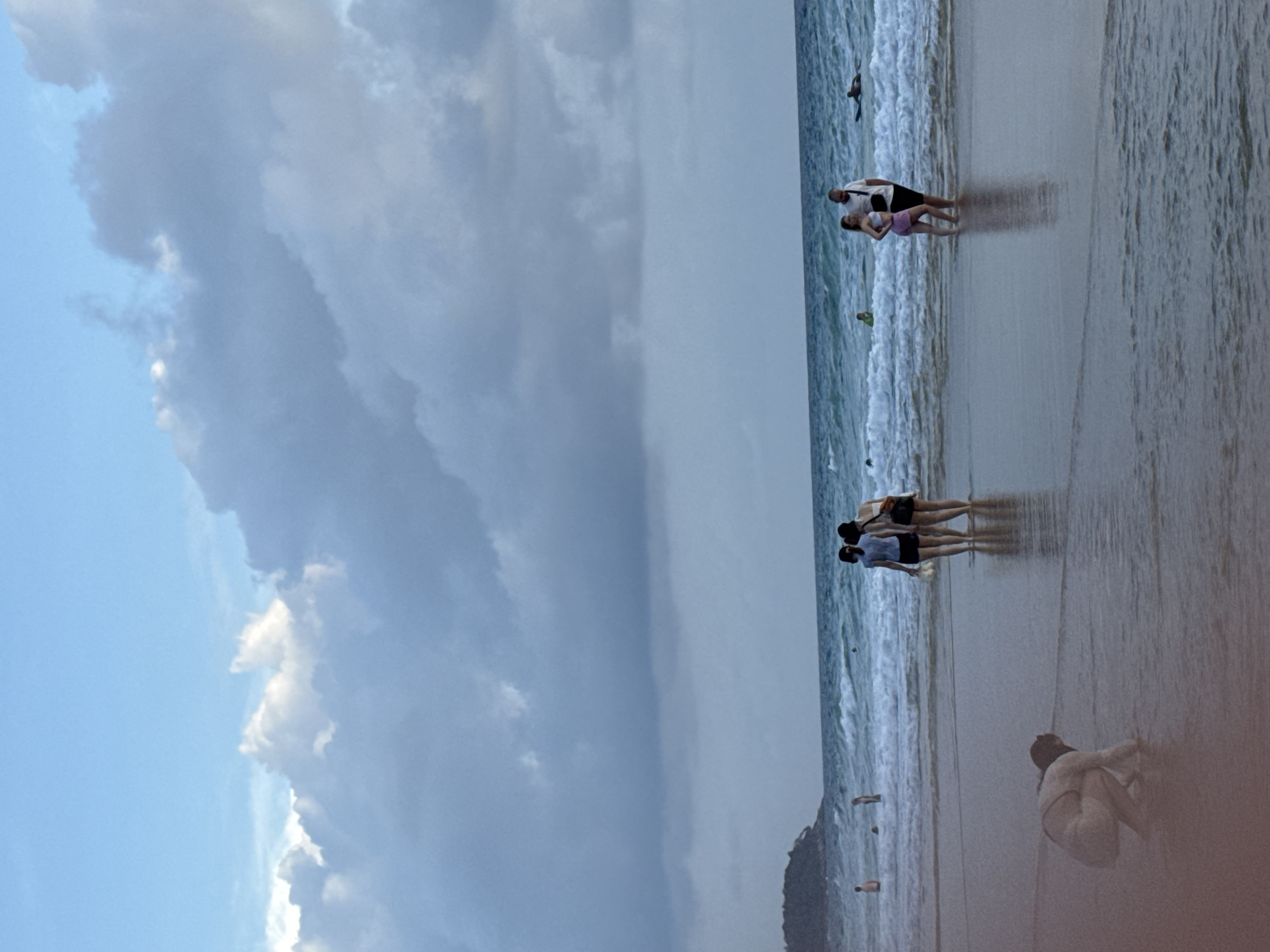 The image depicts a beach scene in Karon, Thailand, under a cloudy sky with patches of blue. In the foreground, a person is crouched down on the wet sand at the edge of the water. Further back, several people are visible. A group of three women stand together, two in the foreground of the group and one slightly behind. To their right, a man and a woman stand in the shallow water, looking at each other. In the background, other individuals are scattered along the beach and in the water, with one person lying on a surfboard. The ocean is active with moderate waves. A distant headland with vegetation is visible on the left side of the image.