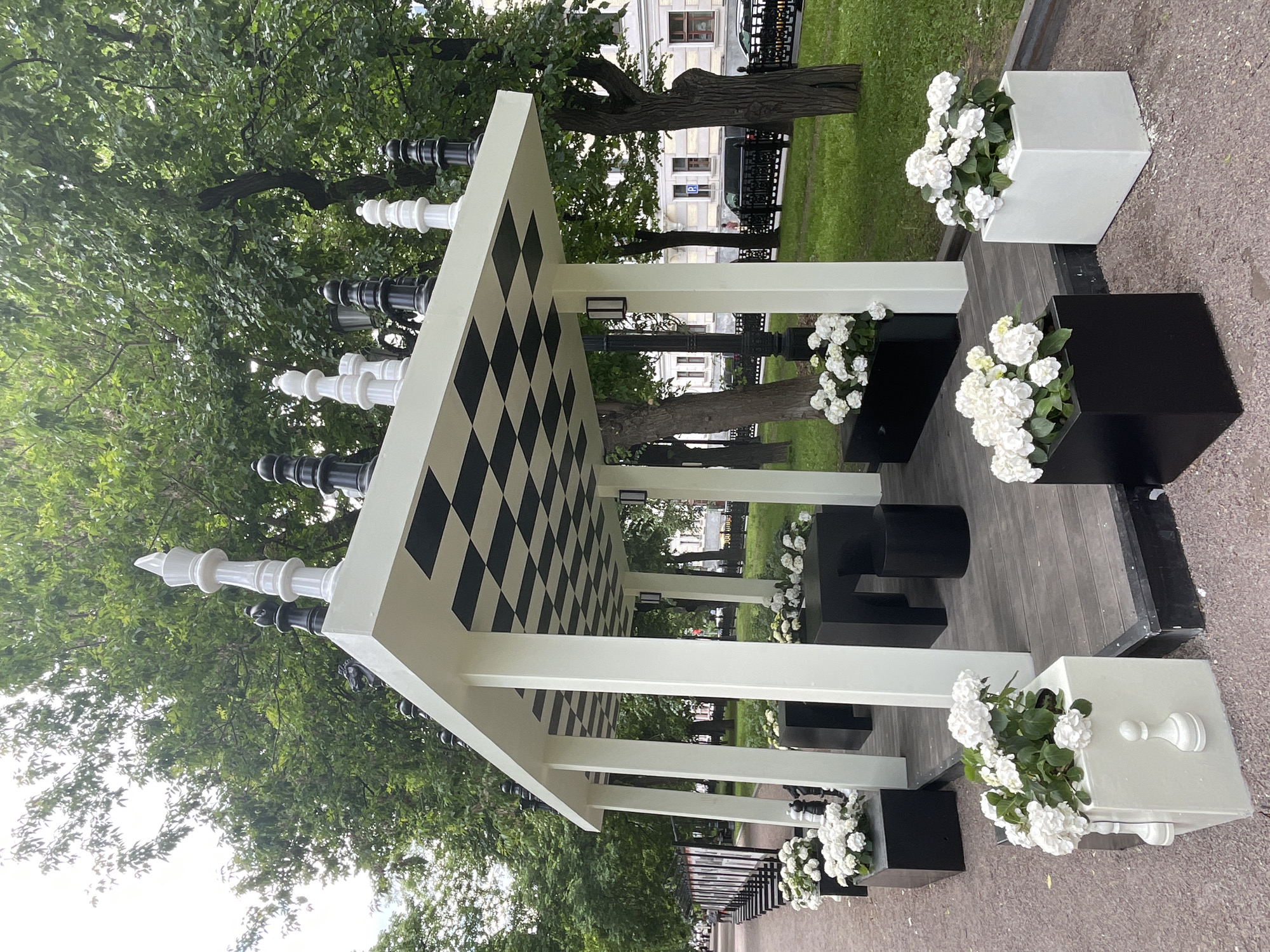 This media file captures a unique outdoor pavilion or pergola, set in a park-like environment in Moscow, Russia, during the daytime, likely under an overcast sky.
The central subject is a large, decorative structure inspired by a chessboard. It features a prominent black and white checkered ceiling supported by sturdy white pillars. Along the top edges of the pavilion, oversized, stylized chess pieces—including kings, queens, rooks, and pawns—are arranged in both white and black, creating a grand, playful design element.
Beneath the pavilion, black benches or seating areas are visible. Complementing the structure's theme, numerous black and white cube-shaped planters are placed around its base. These planters are filled with clusters of white flowers, likely hydrangeas, adding a touch of natural beauty to the monochromatic scheme.
The scene is set outdoors, with abundant green trees and grass surrounding the structure. A paved path or walkway is visible in the foreground and around the pavilion. In the background, beyond the trees, urban elements such as buildings with windows and a dark metal fence can be seen, indicating an urban park setting. No people are present in the image, suggesting a serene moment or an artistic installation.
A notable detail in the distant background, seen on one of the buildings, is a white "P" sign, commonly used to indicate parking. The overall aesthetic is clean, striking, and strongly themed around chess, combining architectural elements with natural landscaping.