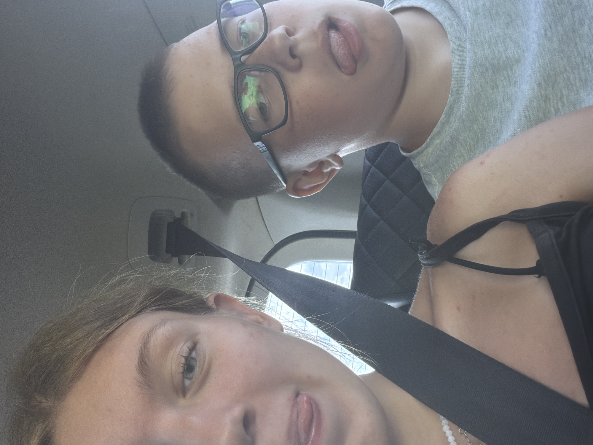 Two individuals are in the back seat of a vehicle, with a black seatbelt prominently displayed across both. On the left, a young female is partially visible, her face turned towards the camera. Her left eye is open, and her tongue is sticking out. She is wearing a black strap top and a white beaded necklace. On the right, a young male wearing glasses is also facing the camera, with his tongue sticking out. He is wearing a light gray t-shirt. The background suggests the interior of a car, with padded seats and a window reflecting light. The image was taken in Novoe Feizullovo, Russia.