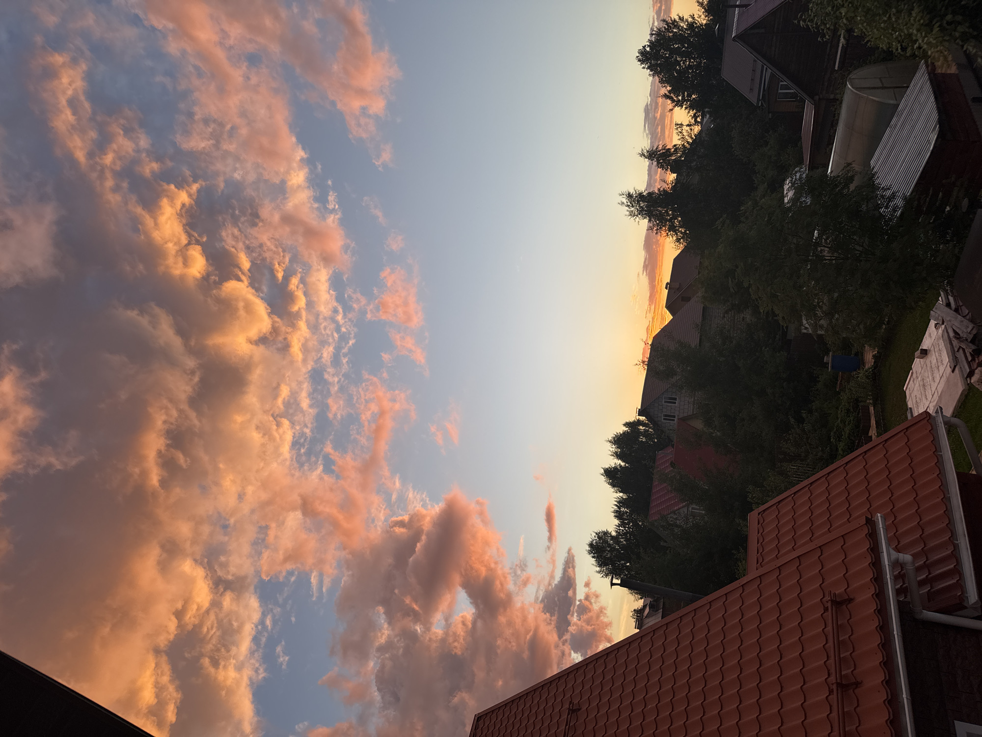 The image captures a dramatic sunset over a residential area, likely in the city of Perm, Russia. The sky is filled with voluminous clouds painted in vibrant hues of orange and pink, illuminated by the setting sun. These clouds are set against a backdrop of a clear, gradient blue sky. In the foreground, the reddish-brown tiled roof of a house juts into the frame from the left, with its gutters and downspout visible. Further into the scene, a cluster of houses with similarly colored roofs and dark green trees are nestled together. The trees obscure much of the lower portion of the houses, but glimpses of chimneys and windows can be seen. A greenhouse and a few other structures are interspersed among the trees and houses, suggesting a semi-rural or suburban setting. The overall impression is one of a peaceful evening scene, with nature's beauty taking center stage.