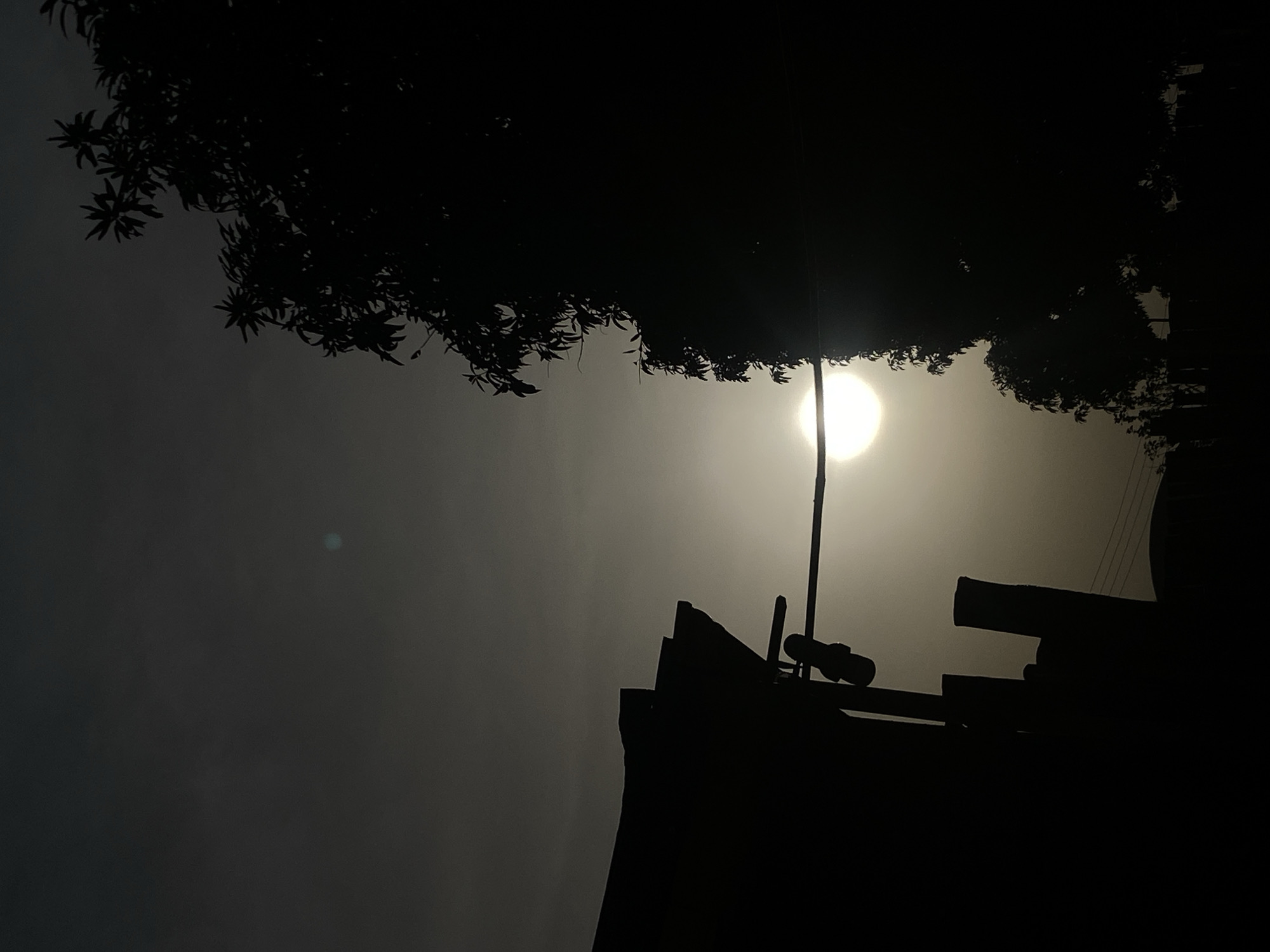 The image shows a silhouette of trees and a rooftop against a bright, hazy sky, likely the sun. A pole extends upward near the center of the frame. The location, Barden Yana, Nigeria, cannot be confirmed based on the visual information.