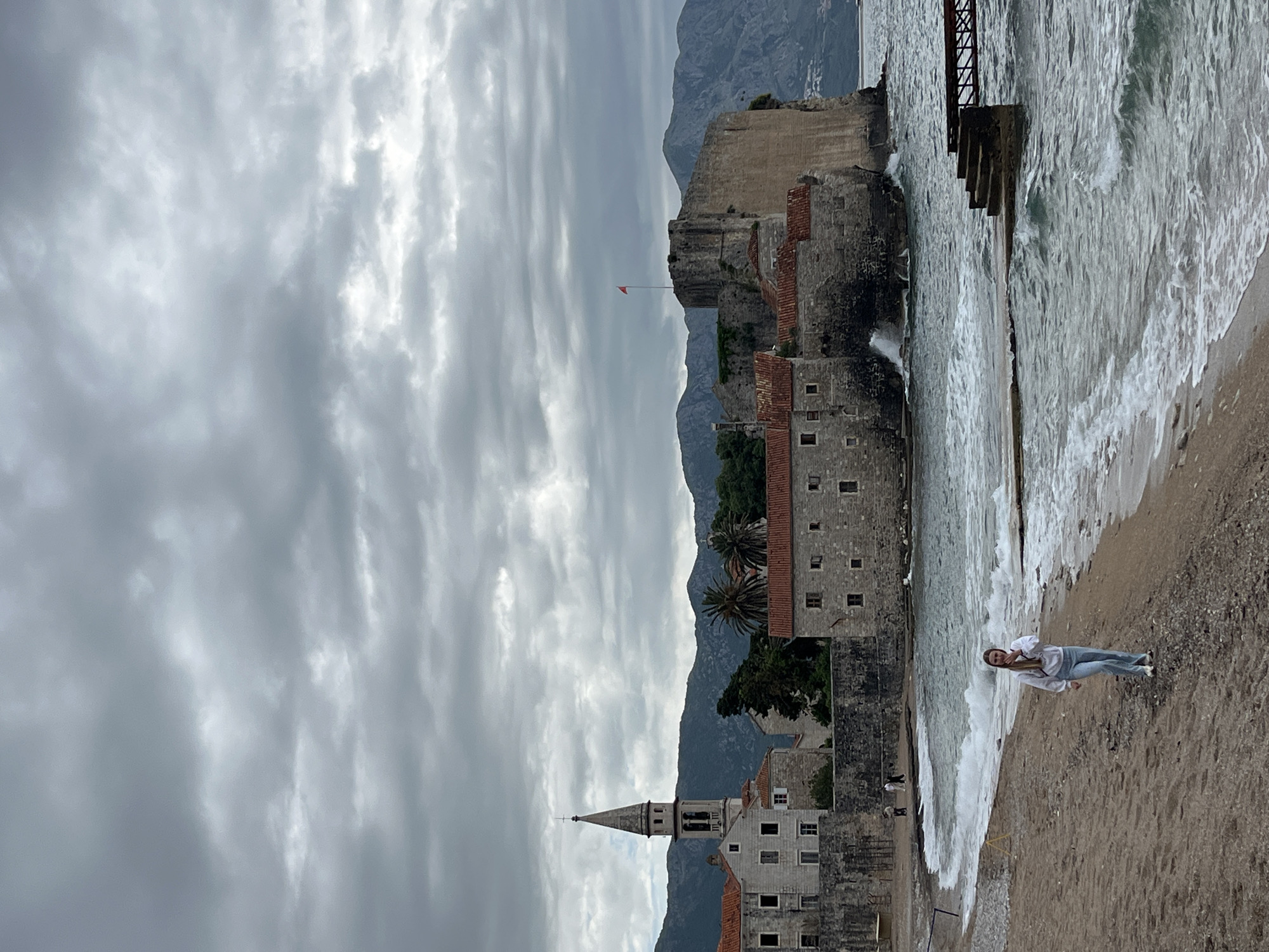 The image shows a cloudy day at the beach near the old town of Budva, Montenegro. The old town's architecture is visible, including stone buildings with red tile roofs, a church spire, and sections of the city walls. Mountains form a backdrop. A woman is seen on the beach, close to the water's edge. Waves are breaking on the shore. Some metal structures or supports are present near the water. The sky is overcast with gray clouds.