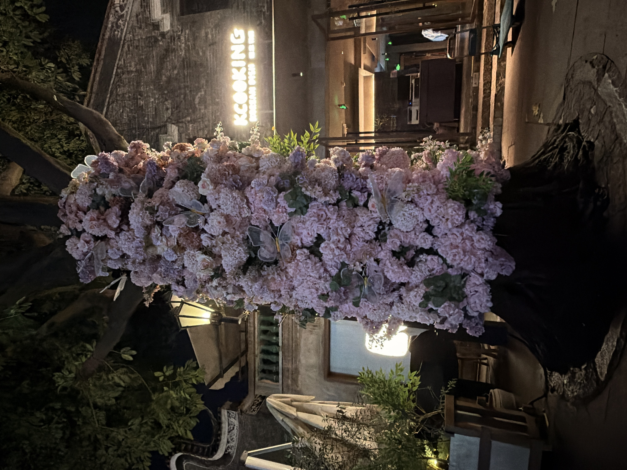 A tree trunk decorated with pink flowers and butterflies stands in front of a building with a neon sign that reads "KCOOKING". The building appears to be a restaurant. The scene is lit by artificial lights, creating a nighttime ambiance. The ground is paved and there are some plants and outdoor seating areas visible. The image is taken from a slightly low angle, looking up at the decorated tree and the building. There are no people clearly visible in the image. The architecture of the building suggests it might be located in a historic area, possibly in Chancheng District, China.