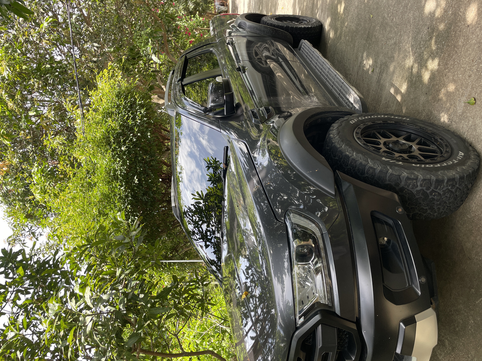 A dark gray Ford Ranger pickup truck is parked on a concrete surface outdoors. The truck is angled towards the viewer, with its front left quarter visible. It has black fender flares, a black grille, and aftermarket black wheels with all-terrain tires. The truck is surrounded by lush green trees and foliage, suggesting a natural or semi-rural environment. The sky is visible through the leaves, appearing bright. The lighting indicates it is daytime, with shadows from the trees cast on the truck and the ground. There are no people or animals present in the image.