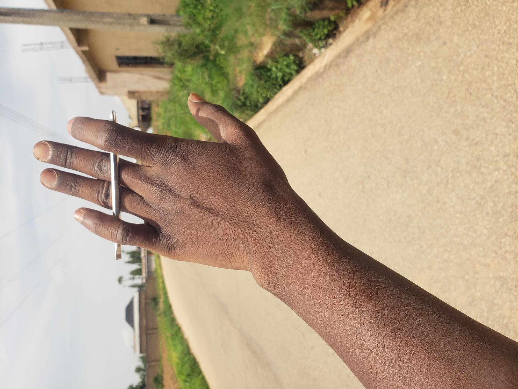 A person's arm and hand are visible in the foreground of the image. The hand is positioned with the palm facing away from the camera, and a silver bar is held between the index and middle fingers. The skin tone of the arm and hand is dark brown. The background depicts a road with a light brown surface, flanked by green vegetation. In the distance, there are buildings, some appearing to be under construction, and trees. The sky is overcast. The scene appears to be in a rural or semi-urban area, possibly on the outskirts of a city like Rayfield, Nigeria, given the architectural styles of the buildings and the surrounding vegetation.