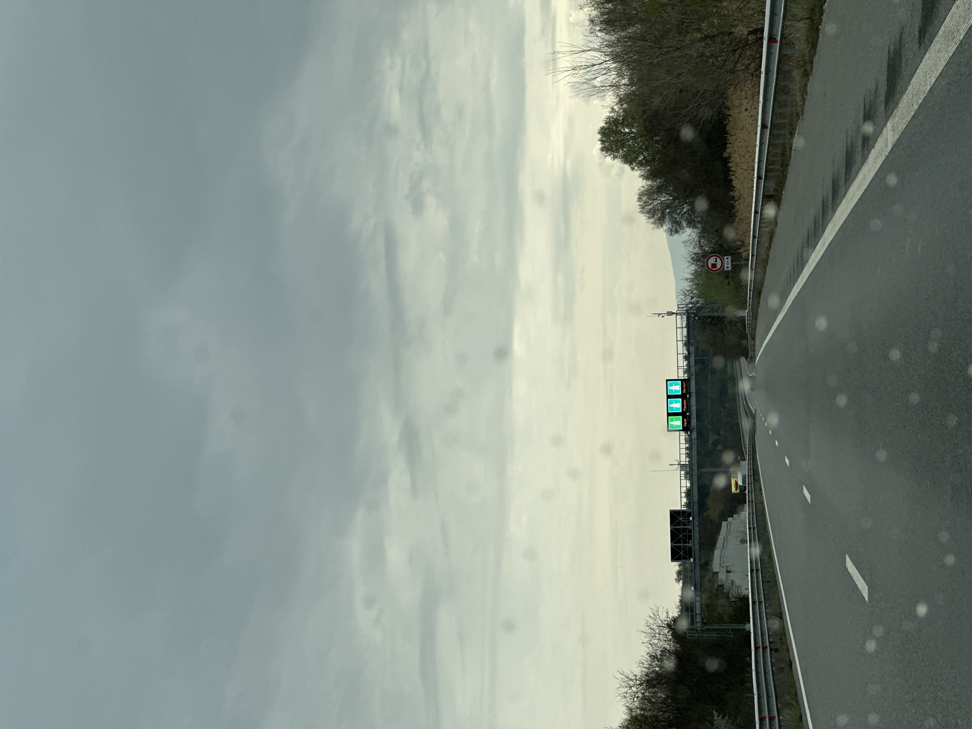 A slightly tilted, long-shot, ground-level image depicts a two-lane highway under a cloudy sky. The highway is wet, presumably from recent rain, and has a white dashed line separating the lanes. To the right of the road, a metal guardrail runs along the shoulder. Beyond the guardrail is a wooded area with trees that have bare branches. Along the shoulder, there is a round traffic sign with a red border. Further down the road, an electronic sign displays messages in green. Behind the sign is a bridge structure.
The location in Valkovo, Bulgaria cannot be verified.