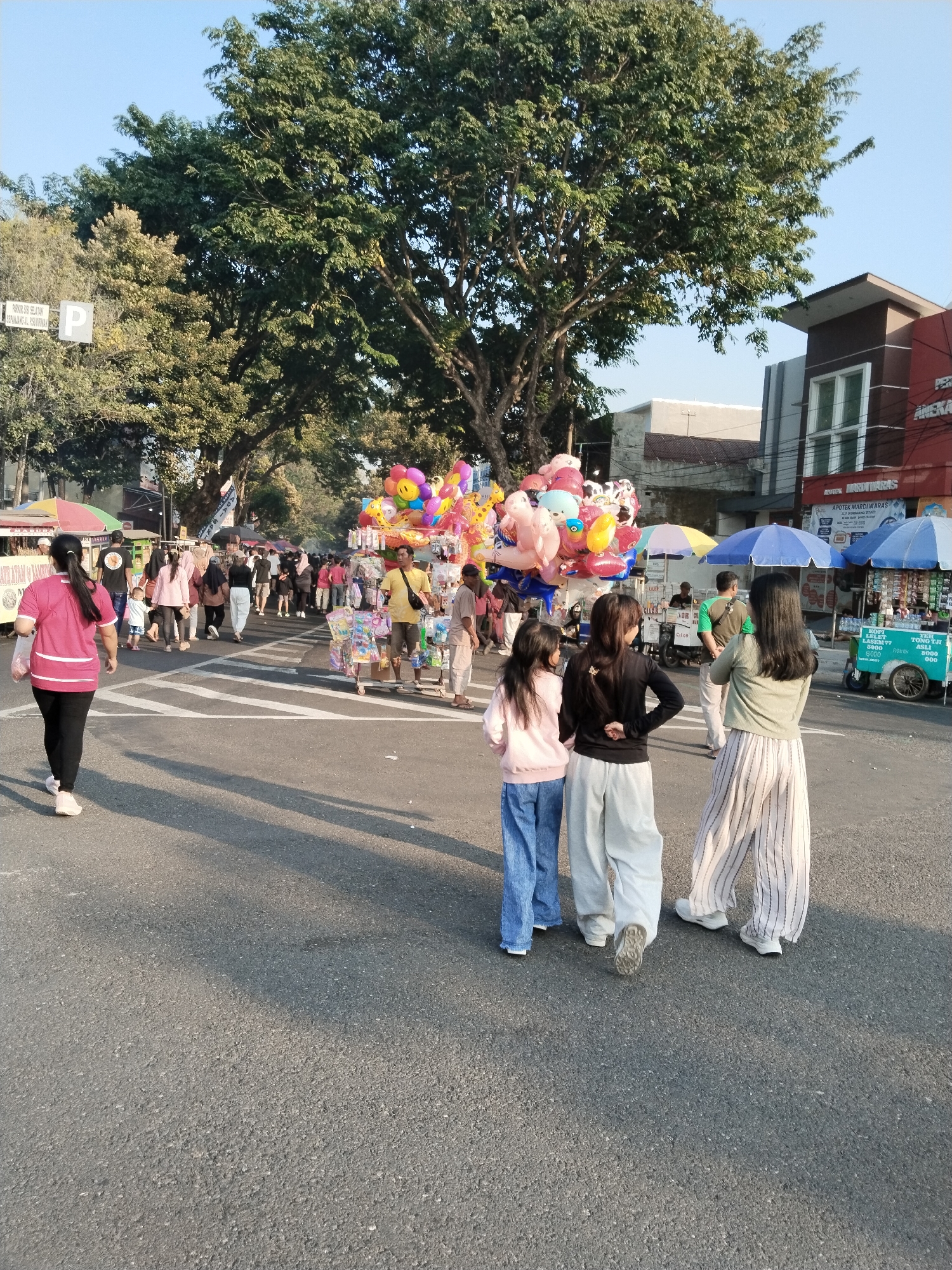 In Kecamatan Pati, Indonesia, a bustling street scene unfolds under a clear sky. A pedestrian street is filled with people, stalls, and a large tree. In the foreground, three individuals walk away from the camera, two women in casual attire and a young girl. Behind them, more people are visible, some walking and others browsing at various stalls selling balloons, food, and other goods. The street is lined with trees and buildings, including a pharmacy. The atmosphere appears lively and social, with people enjoying a day out.
