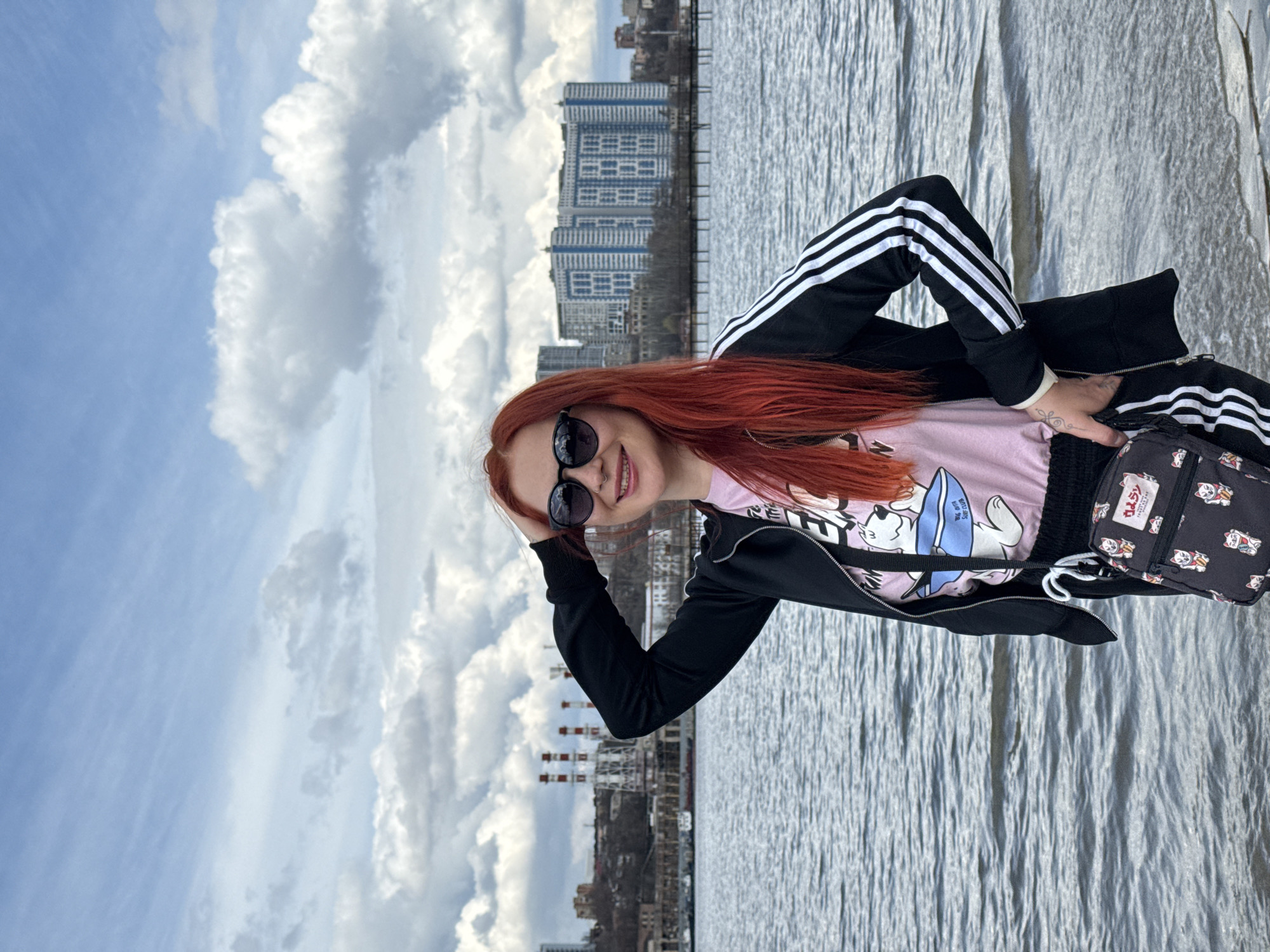 A woman with red hair and sunglasses is standing next to a body of water with tall buildings visible in the background. She is wearing a pink shirt with a graphic design, a black jacket with white stripes on the sleeves, and a black bag with a pattern of cat figures on it. She is holding her sunglasses with her left hand and smiling at the camera. The sky is blue with puffy white clouds. The photo was taken in Rostov-on-Don, Russia, near the embankment of the Don River. In the background, there are industrial chimneys and the city's skyline. The water is calm with small waves. The image is rotated 90 degrees clockwise.
