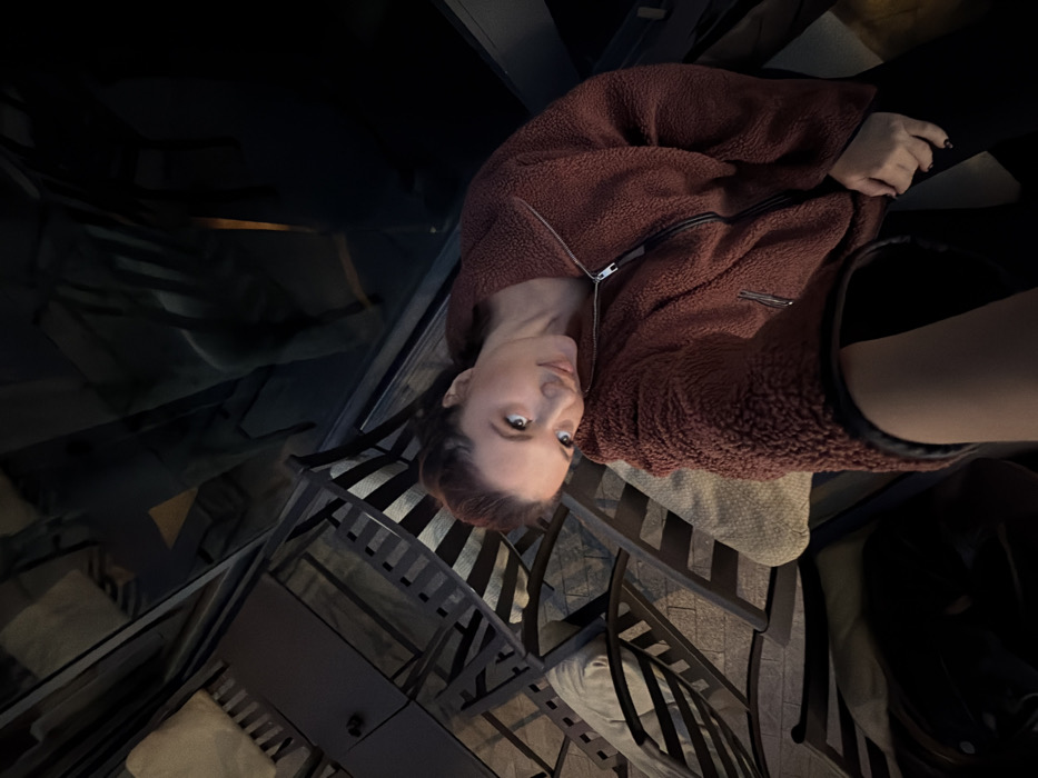 A female individual, appearing to be in her 20s or 30s, is depicted in an angled, head-and-torso self-portrait taken within a commercial establishment in Belgrade, Serbia. She has light-colored eyes and reddish-brown hair pulled back. She is wearing a rust-colored, long-sleeved fleece jacket with a silver zipper, worn over dark trousers. Her left arm is extended towards the camera, while her right hand rests on her thigh. She maintains direct eye contact with a slight smile, her head tilted slightly to the right. Her fingernails are painted dark.
The surrounding environment is dimly lit, suggesting evening or night. The background features multiple dark-framed chairs with slatted backs and seats, some fitted with light-colored, possibly beige or cream, cushions. To the left of the subject, several chairs are stacked on tables, indicating a section of the establishment that is closed, being cleaned, or arranged for storage. The floor beneath the furniture appears to be paved with light-colored, rectangular blocks or tiles. To the right, a dark, reflective surface, likely a window or glass partition, shows indistinct reflections of the interior or external environment. The overall scene suggests an indoor or sheltered outdoor seating area of a cafe or restaurant.