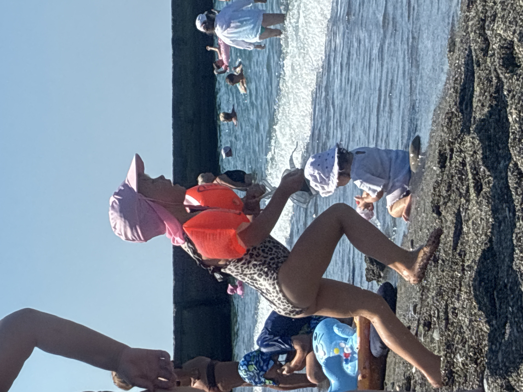 The image shows a group of people at the beach in Sochi, Russia. In the foreground, a young girl is sitting on the shore, wearing a leopard-print swimsuit, a pink hat, and an orange life jacket. She is holding a clear plastic cup and appears to be playing with it. To her left, another person's arm is visible, and to her right, a blue inflatable toy with a cartoon character is present.
In the background, several other people are visible in the shallow water and along the shoreline. Some are wading, while others appear to be playing or swimming. The water is calm and blue, with gentle waves lapping at the shore. The sun is shining brightly, indicating a clear, sunny day, likely in the late morning or early afternoon given the quality of light. The setting is a rocky beach, with dark, coarse pebbles dominating the foreground. Beyond the beach, a dark, elevated structure, possibly a seawall or pier, is visible. The overall scene conveys a sense of a pleasant summer day spent at the beach, with families enjoying outdoor activities. No text is visible in the image.